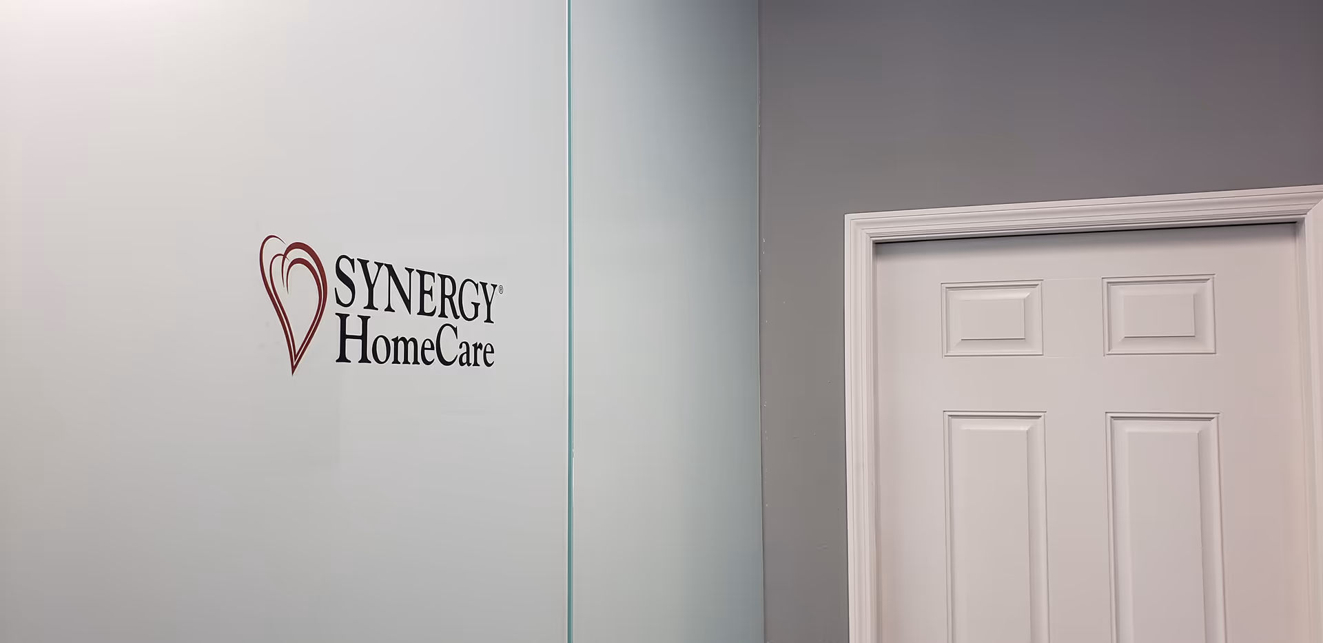 Interior wall area with Synergy HomeCare logo on glass and a white door.