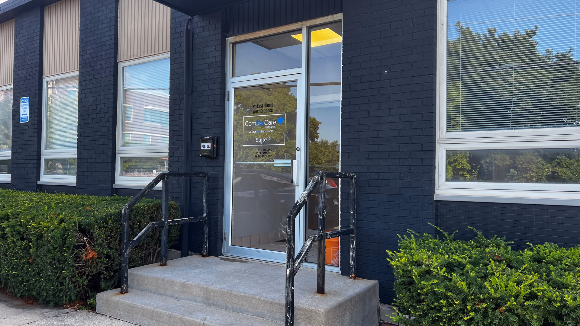 Brick building exterior showing a glass entrance door, concrete steps, and surrounding green shrubs.