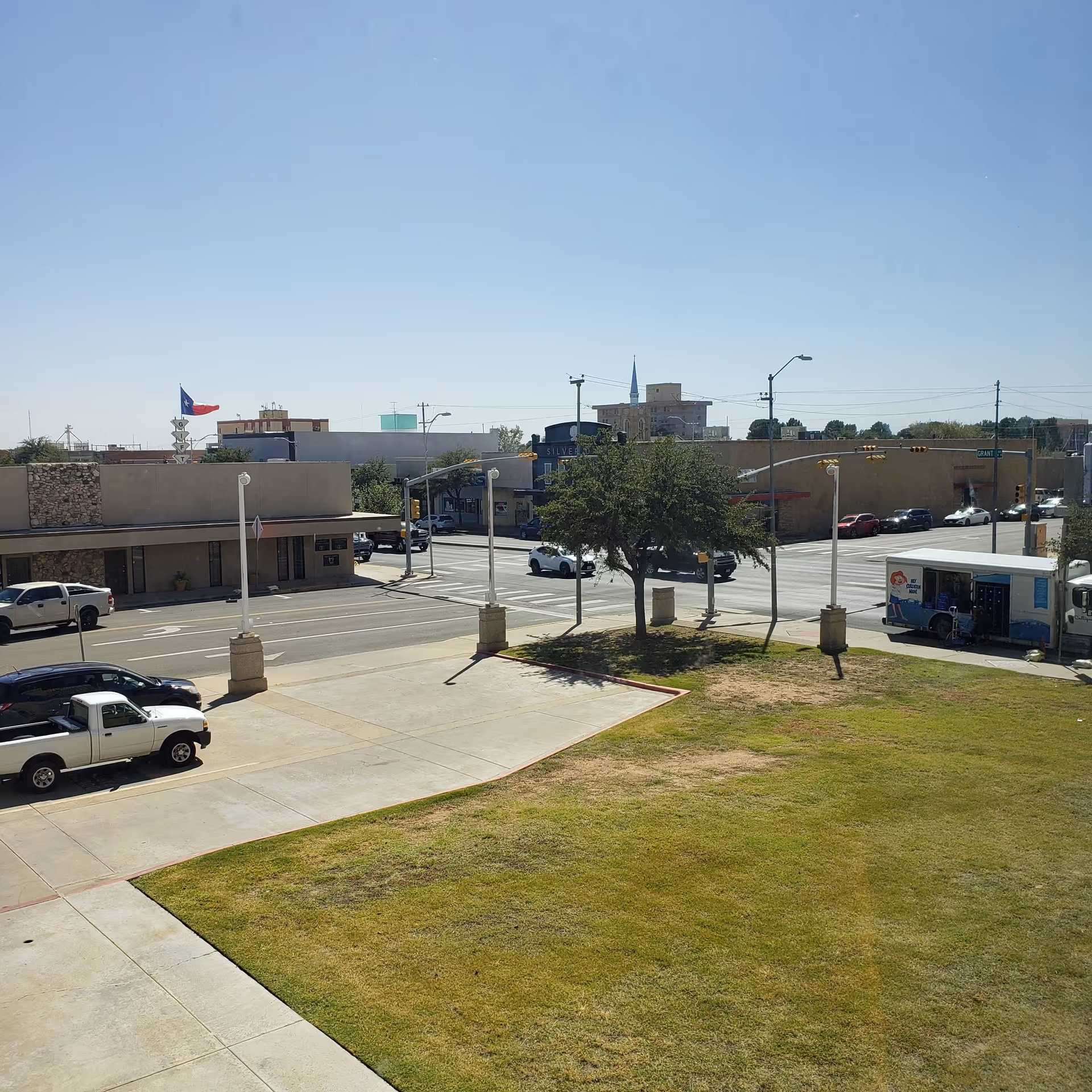 Outdoor street view with a grassy area, trees, parked cars, and surrounding storefronts.