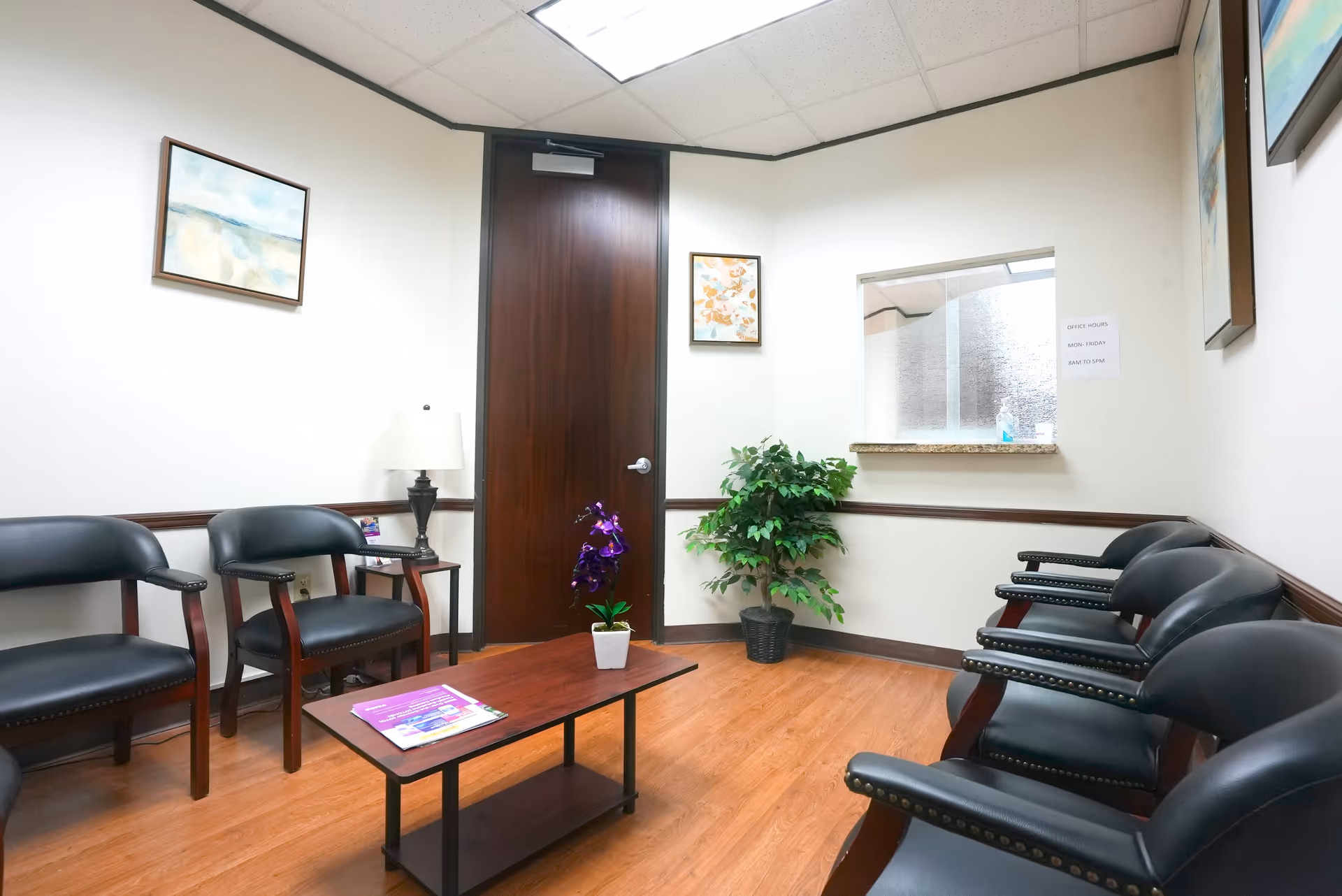 A tidy medical center waiting area with black chairs, a coffee table, and potted plants.