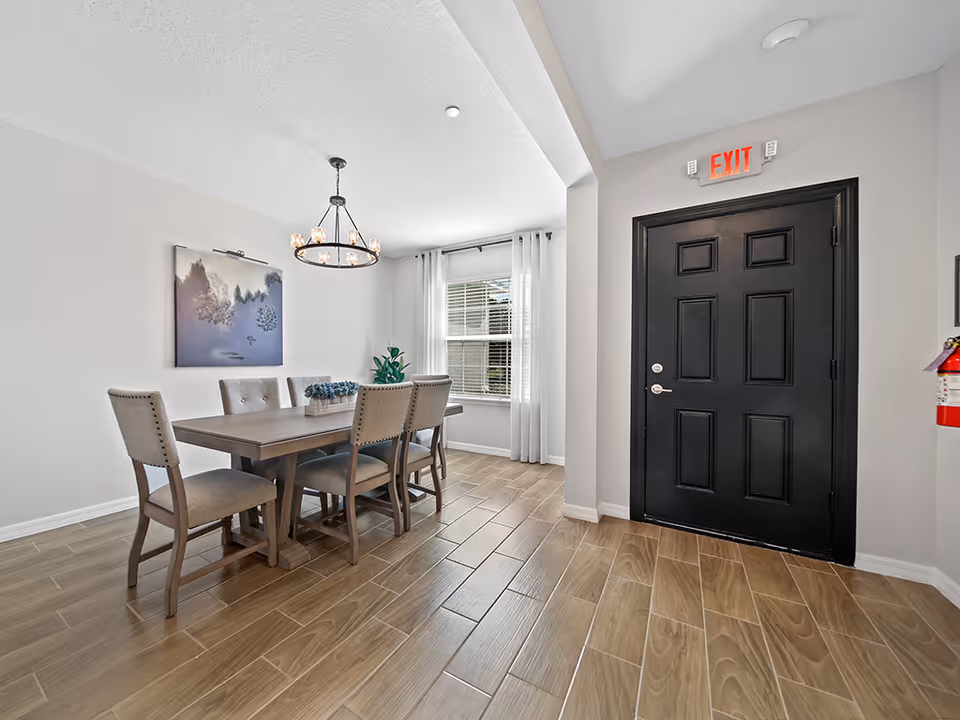 Dining room with wooden table, six upholstered chairs, chandelier, window, and black exit door.
