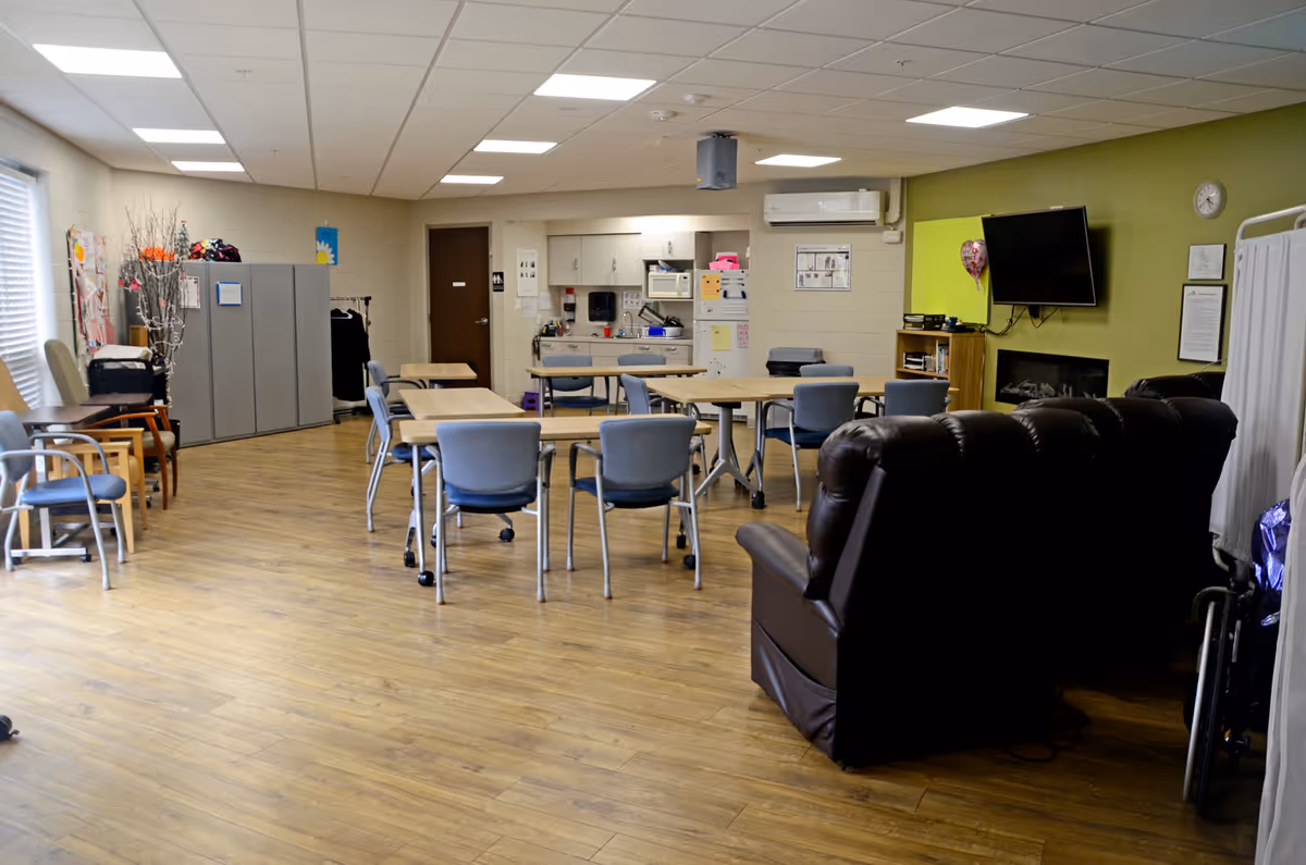 Spacious communal living room with tables, blue chairs, a brown recliner, and wall-mounted TV.