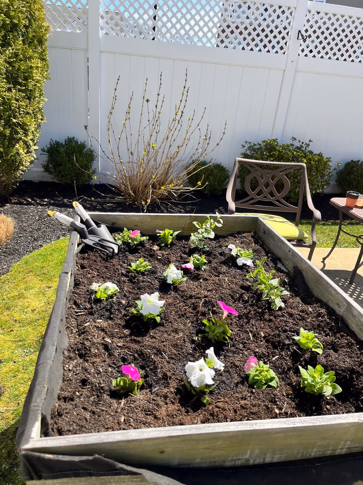 Raised garden bed with flowers, gardening tools, a chair, and a table on a sunny patio.