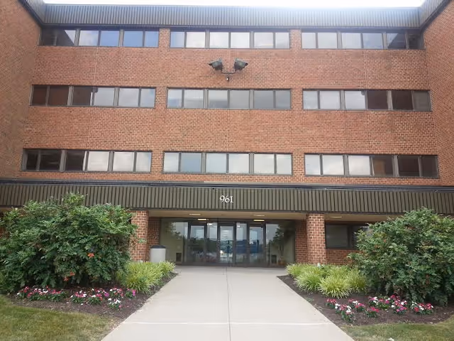 Brick four-story senior living building with glass entrance and landscaped walkway.