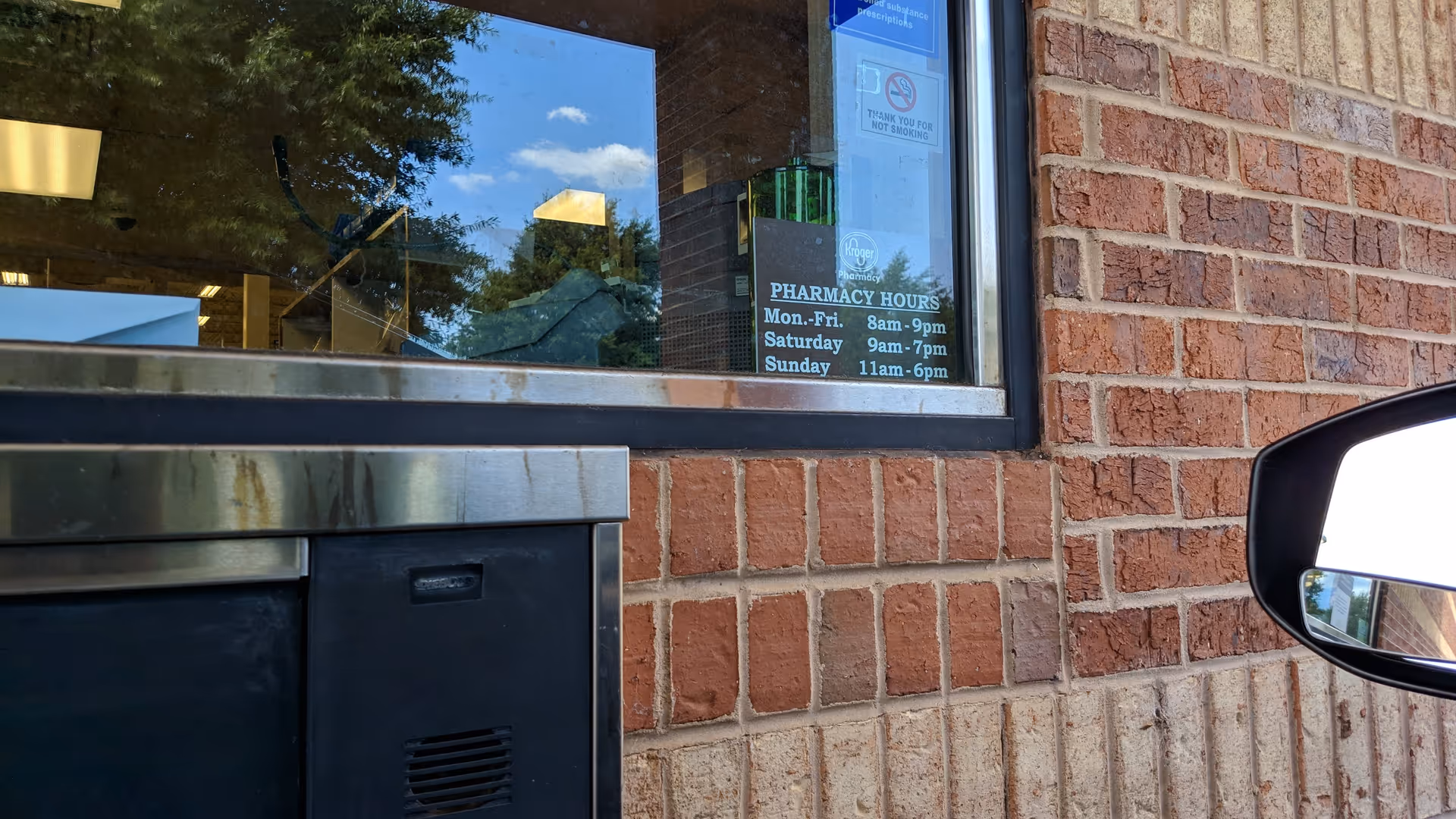 Brick exterior with Kroger Pharmacy window sign and a visible car mirror.