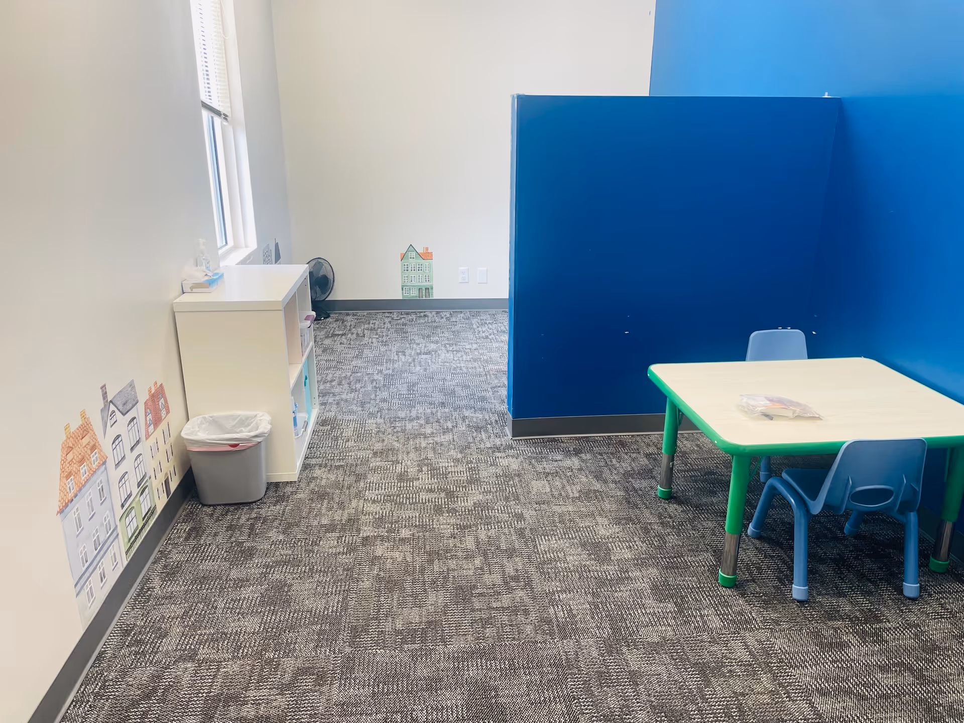 Indoor play area with blue divider, kid-sized table and chairs on a carpeted floor.