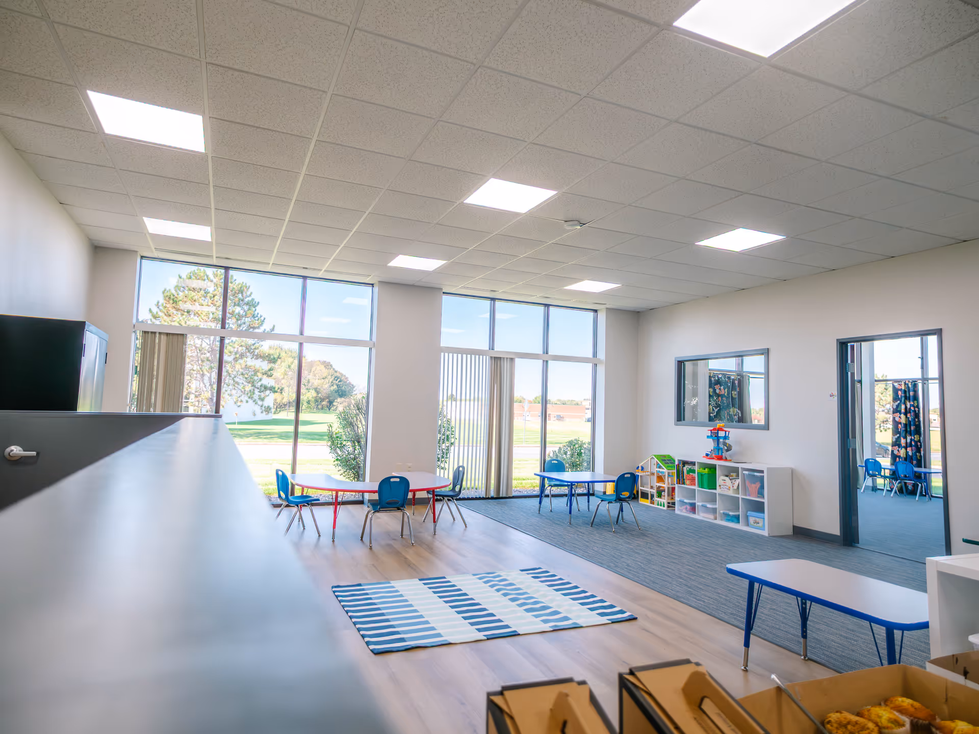 Bright children's activity room with tables, chairs, shelves, and large windows.