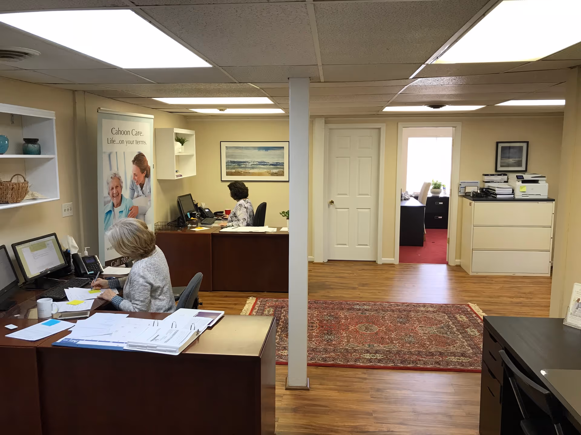 Office workspace with desks, computer stations, a Cahoon Care banner, and two staff members.