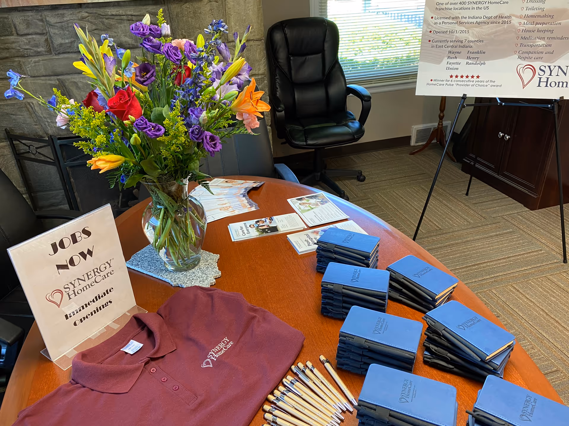Round wooden table with floral arrangement, blue booklets, pens, and a recruitment sign inside a lobby.