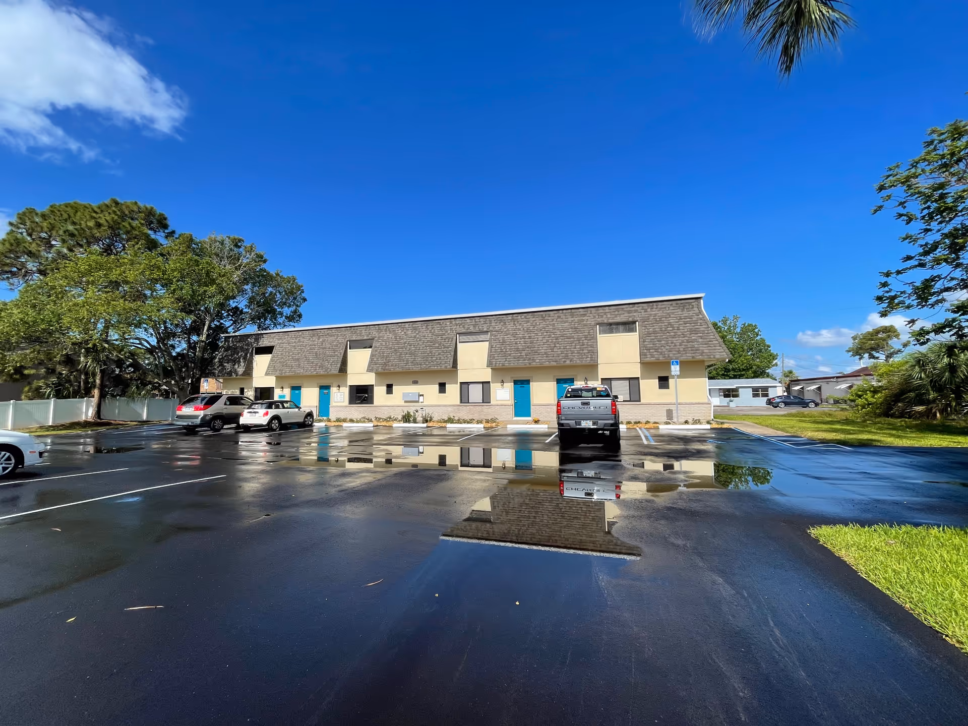Long beige senior care building with blue doors and a parking lot under blue sky.