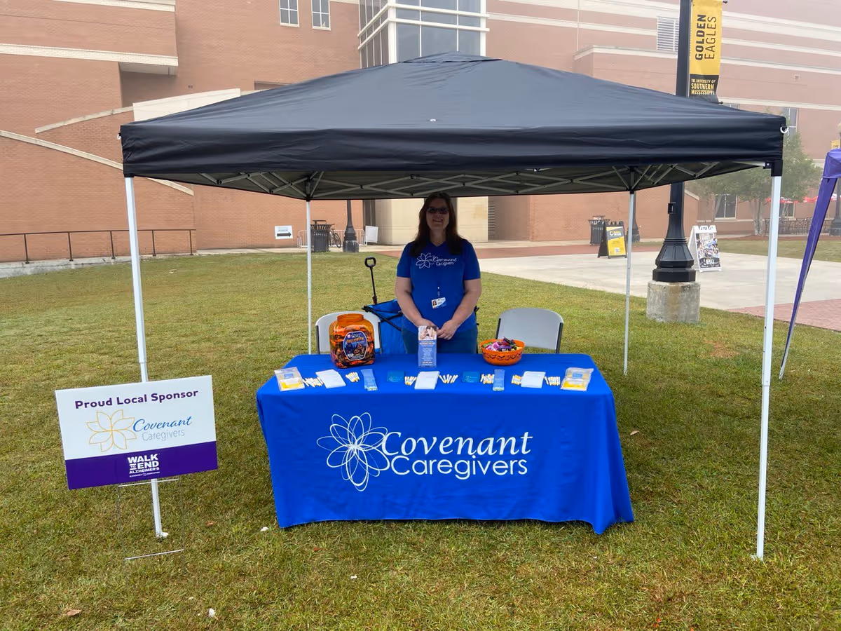 A woman standing behind a Covenant Caregivers table under a black canopy outdoors.
