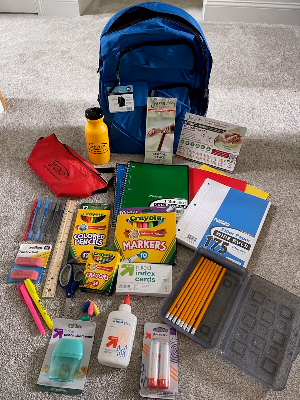 Blue backpack and colorful school supplies spread on carpeted floor in Sahara Home Care facility.