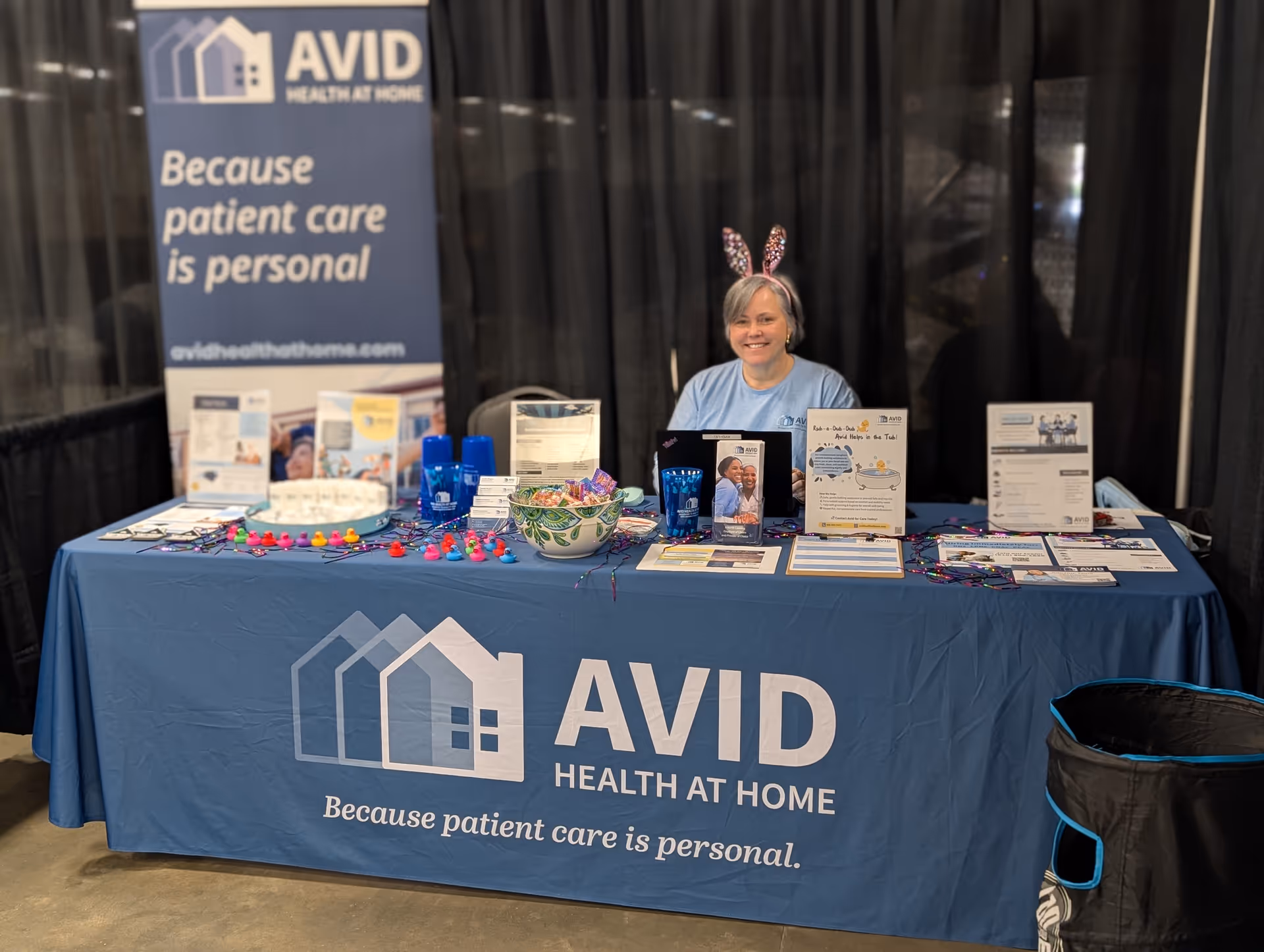 A smiling woman sits behind a promotional booth for Avid Health at Home with brochures.