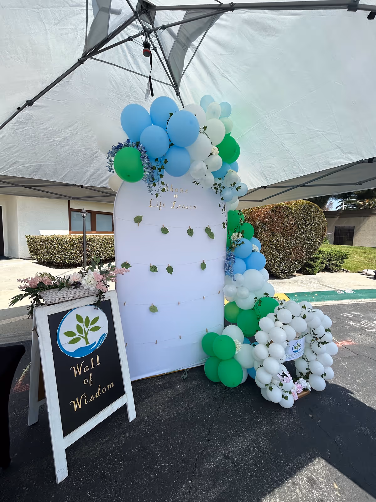 Outdoor event setup with a white arch, blue-green balloons, and a Wall of Wisdom sign.