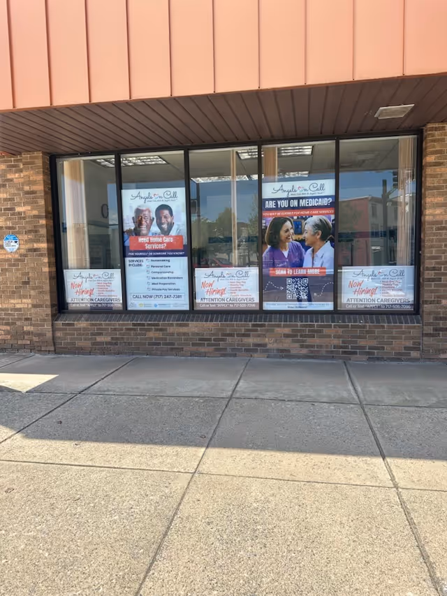 Storefront window displays Angels On Call Home Care posters in a brick building.