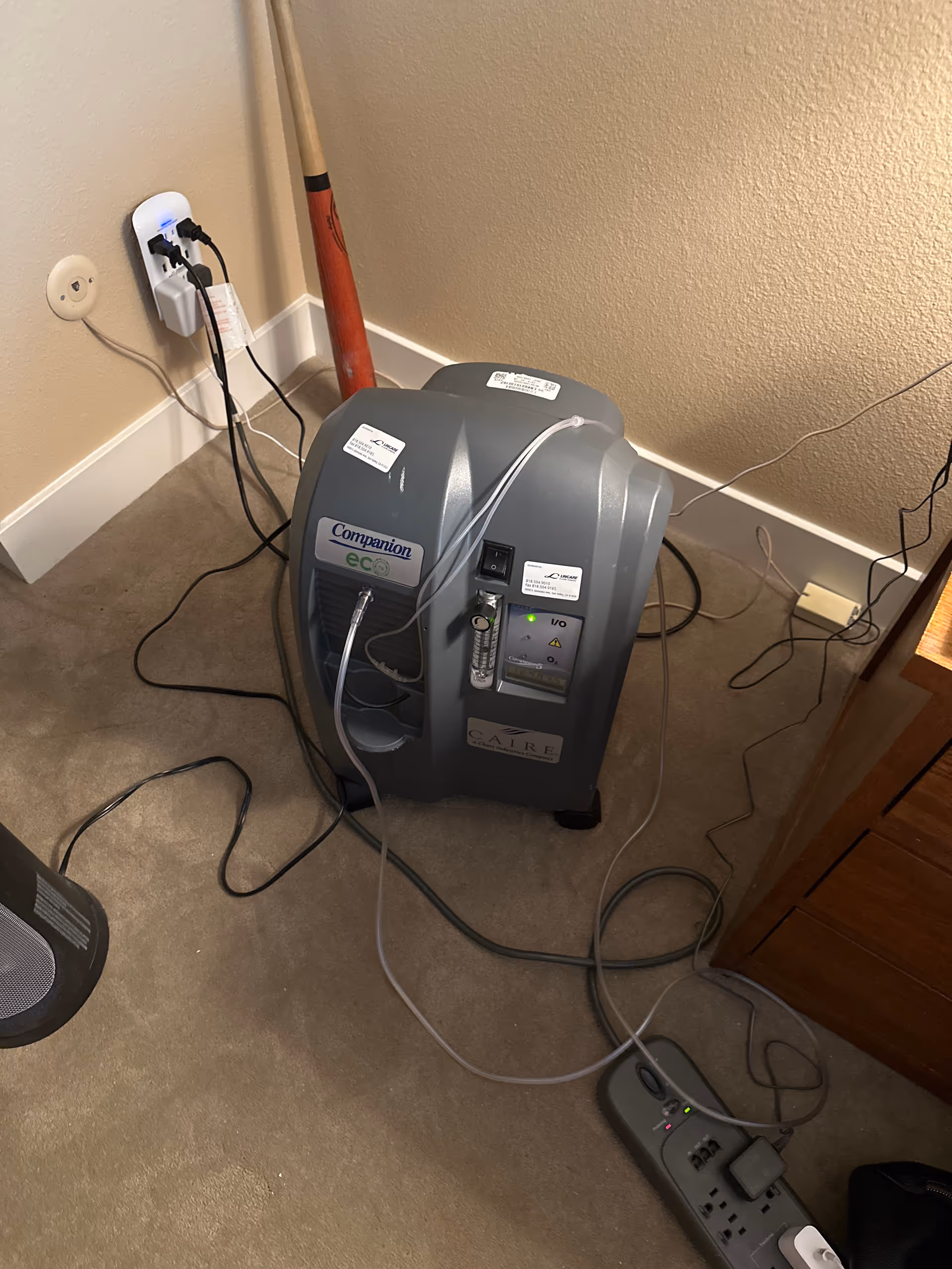 A compact oxygen concentrator sits in a corner with cords plugged into a wall outlet.