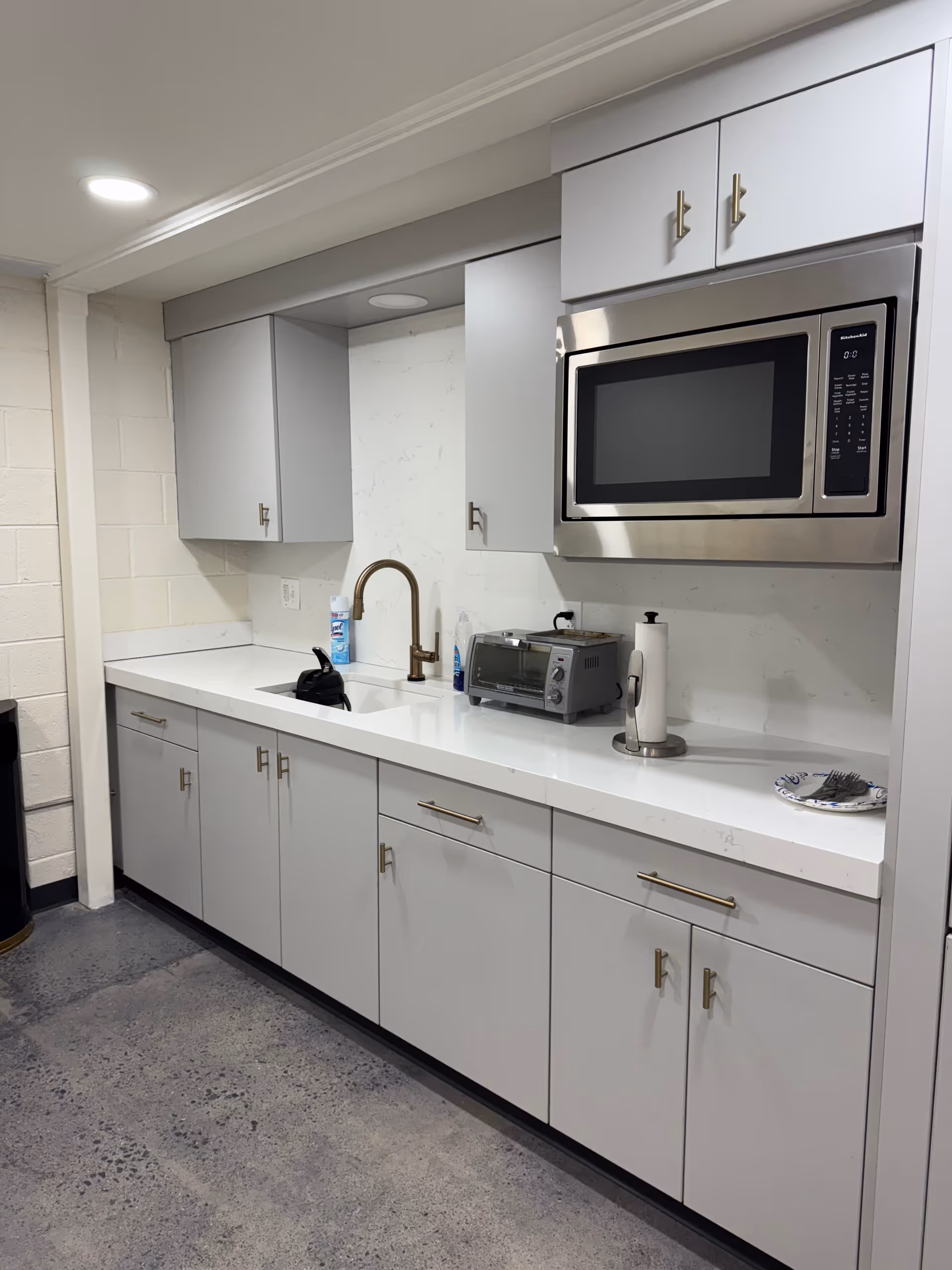 Grey kitchen cabinets surround a sink, microwave, and toaster in a facility kitchenette.