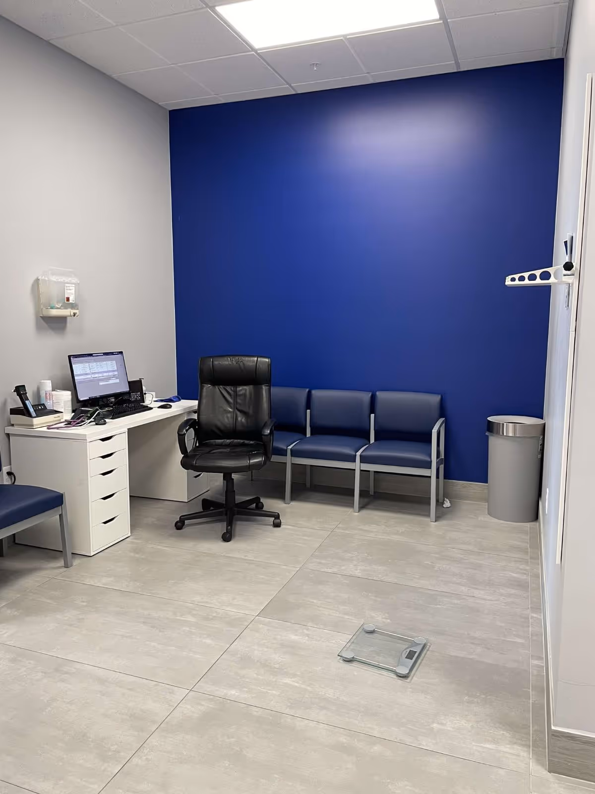 Blue-walled clinic office with desk, computer, a black chair, and blue seating along the wall.