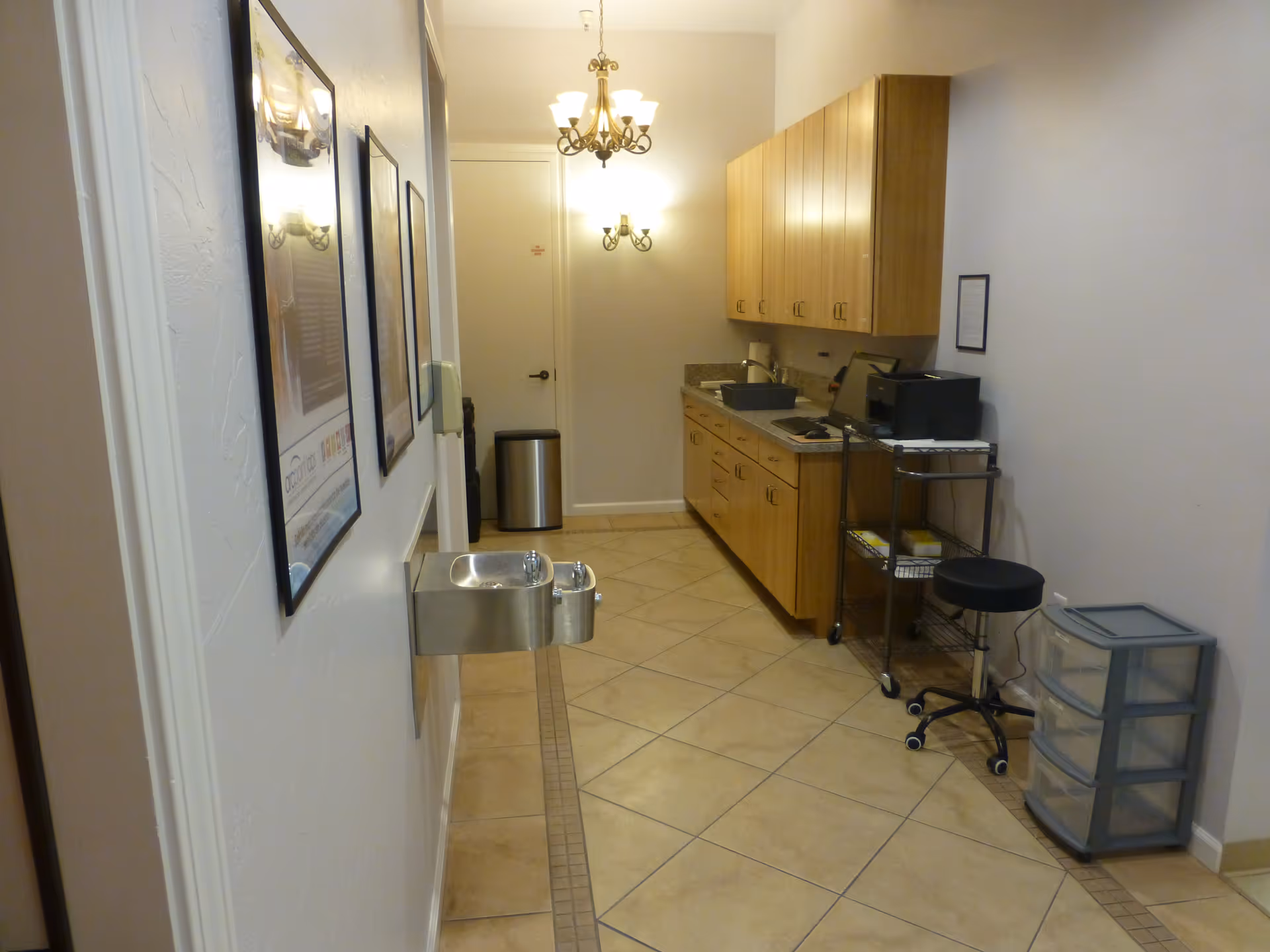 Narrow facility kitchenette with wooden cabinets, sink, counter, wall-mounted water fountain, and rolling printer cart.
