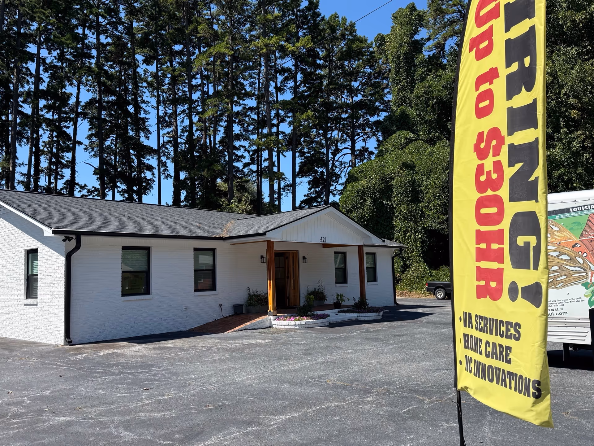 Single-story white building with a covered entrance, a large yellow banner, and tall pine trees behind.