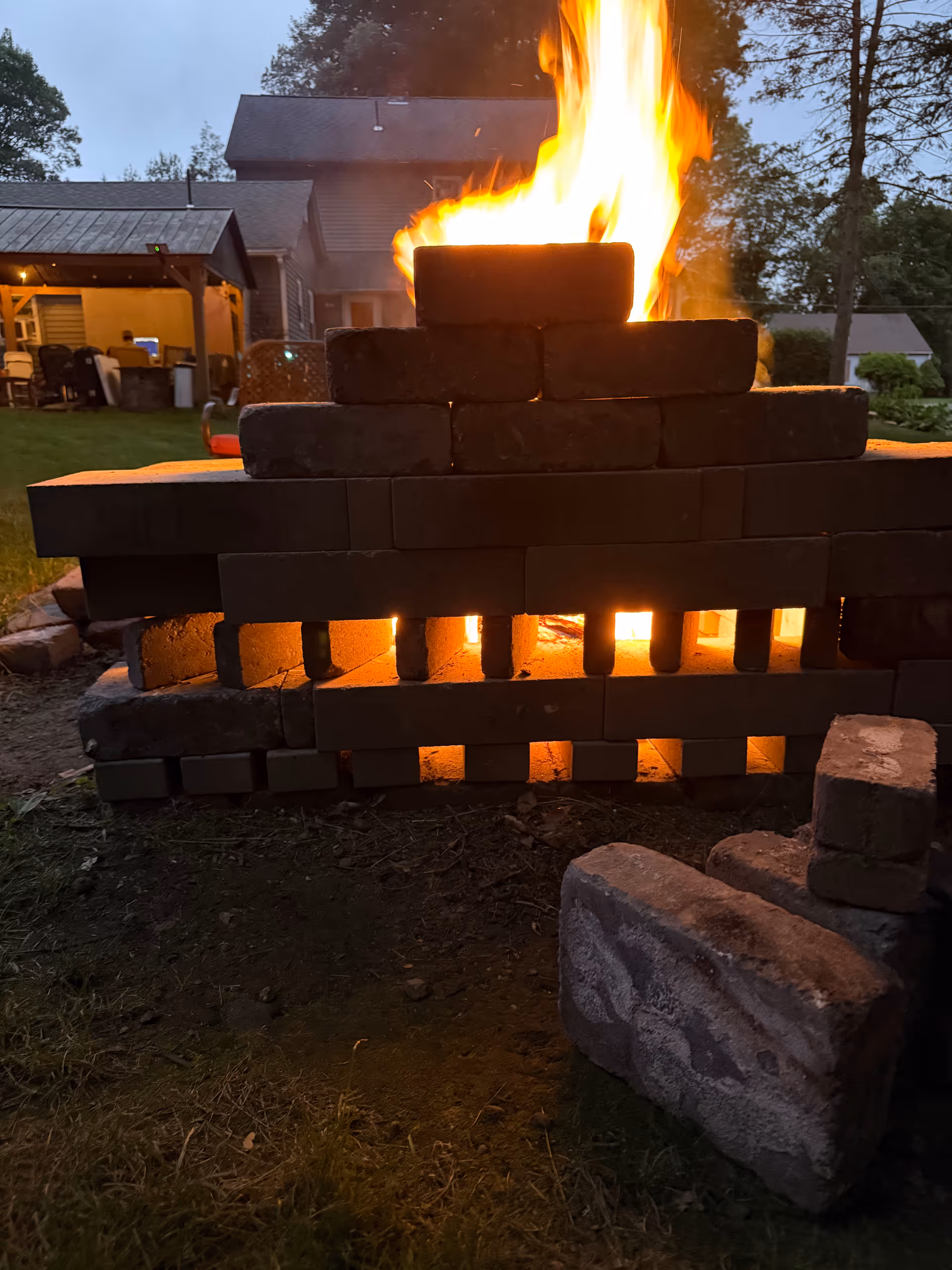 Outdoor brick fire pit with tall flames; background building and patio seating.