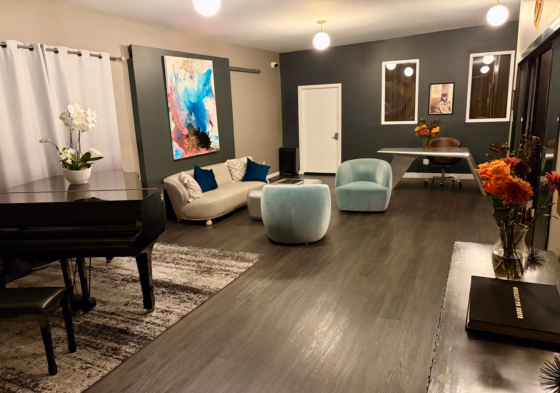 Modern lounge area with piano, teal chairs, sofa, desk, and colorful abstract art.