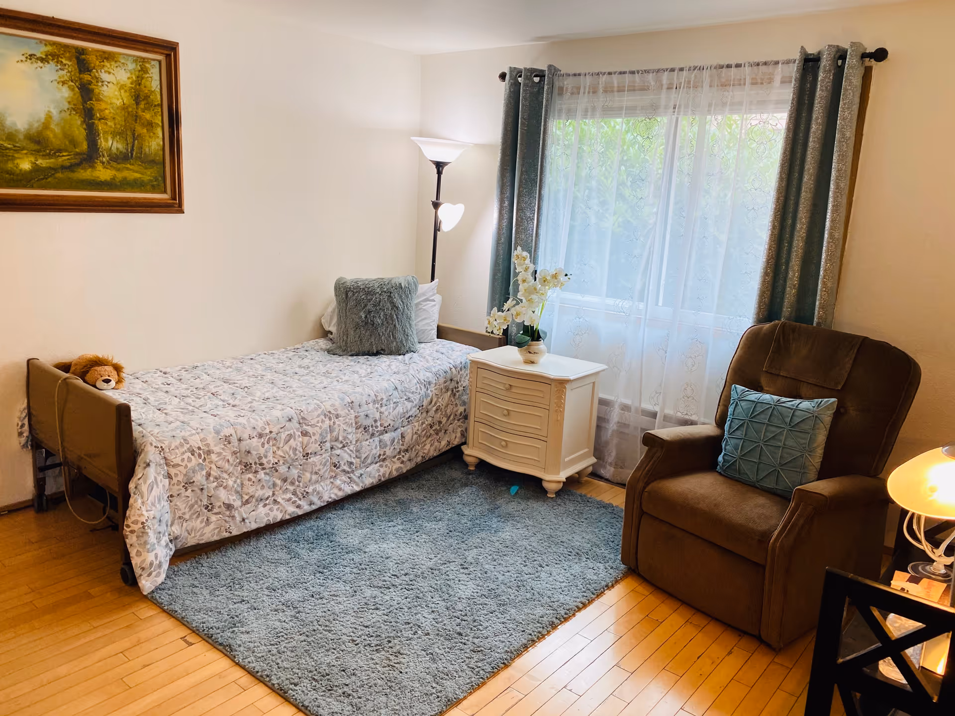 Cozy bedroom in a senior facility with a single bed, nightstand, recliner, and window.