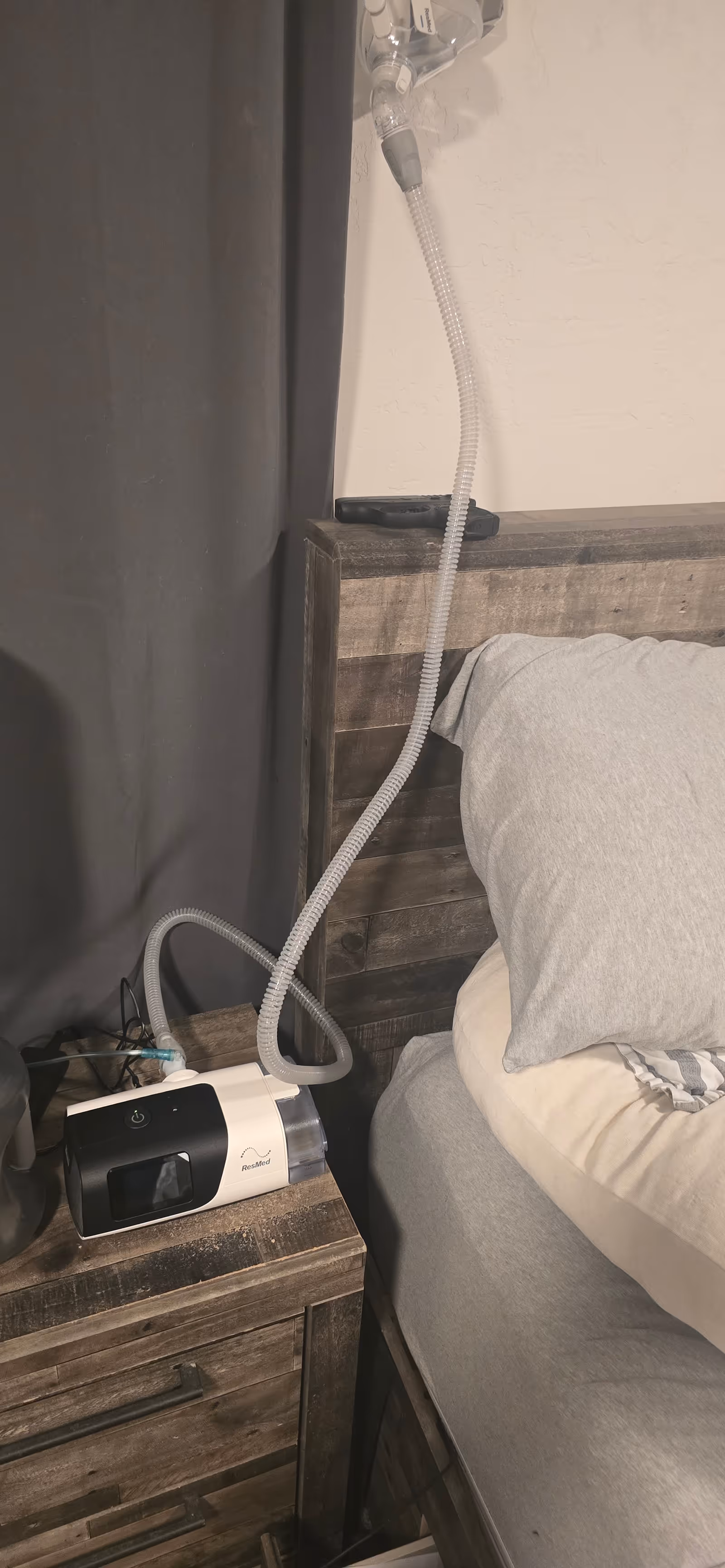 A CPAP machine sits on a wooden nightstand beside a bed with a connected hose.