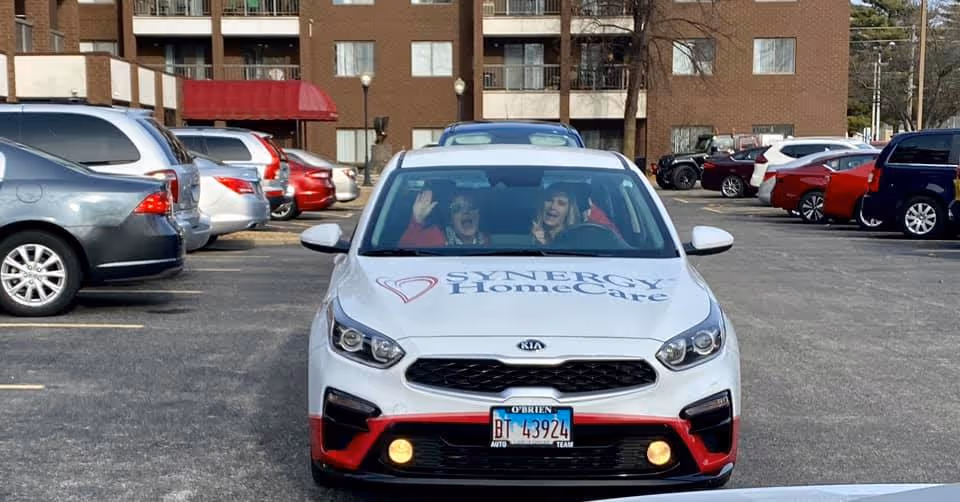 White SYNERGY HomeCare car in a parking lot, two people waving inside, brick building behind.