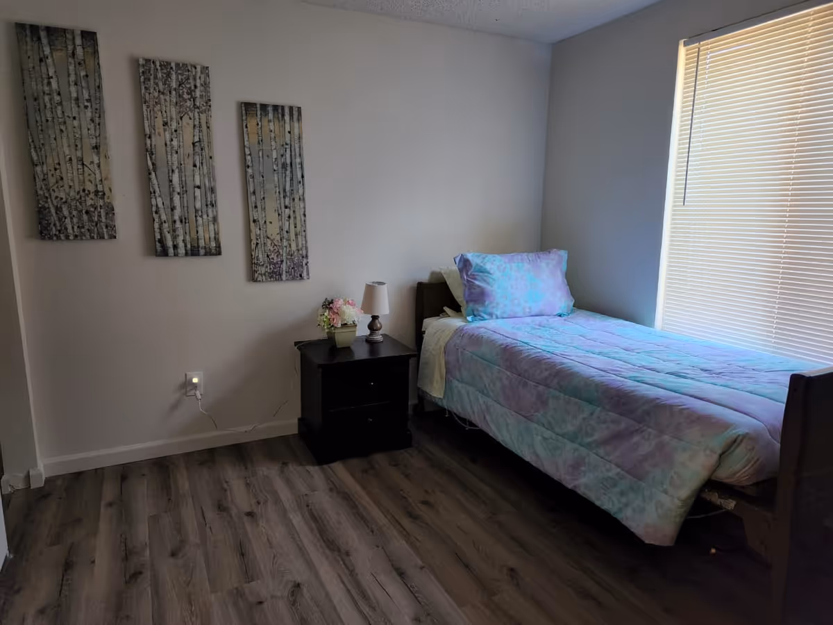 A single bedroom in Blissful Foster Home with bed, nightstand, lamp, blinds.