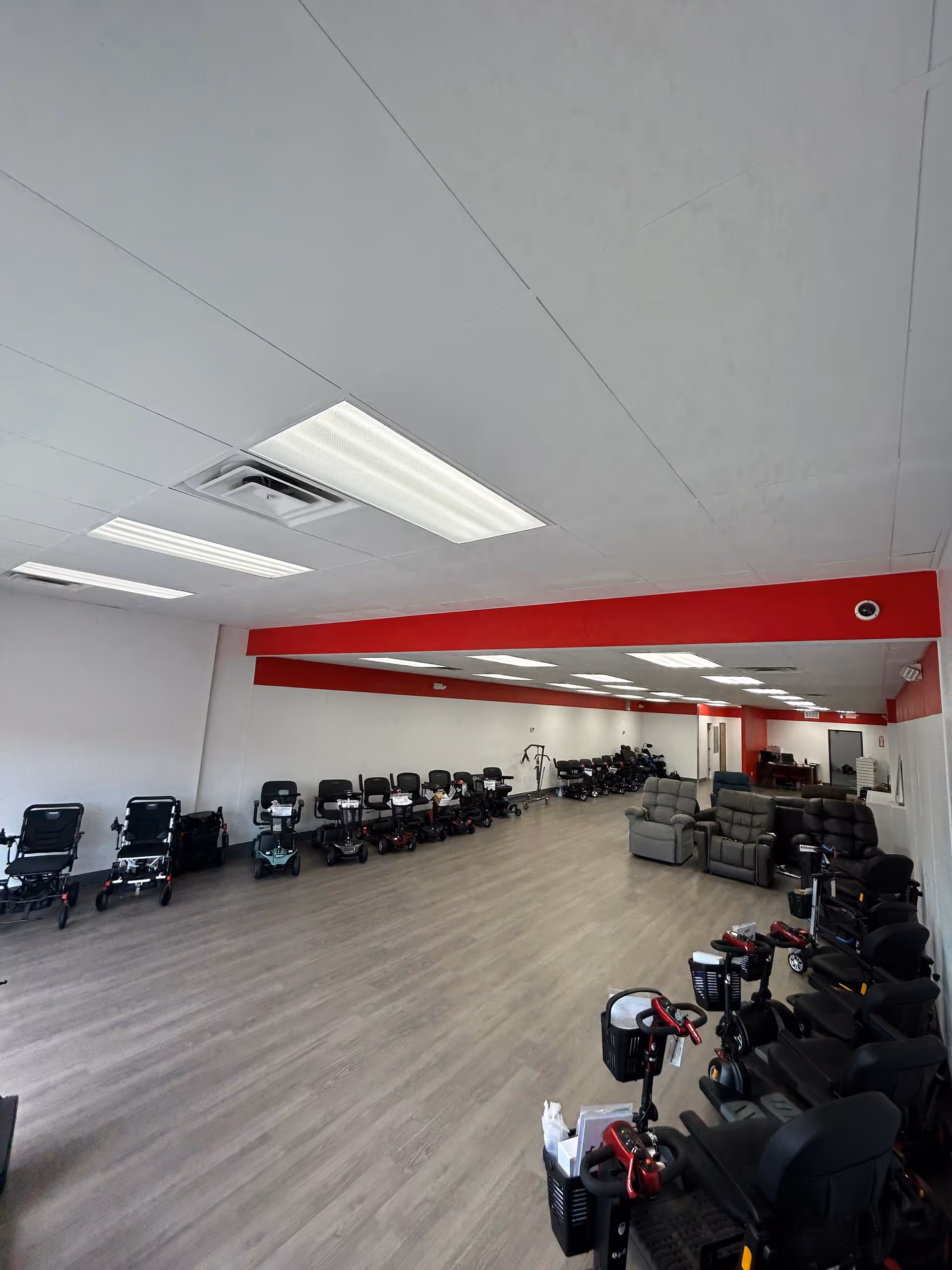 A long room displaying numerous mobility scooters and chairs along a red-accented wall.
