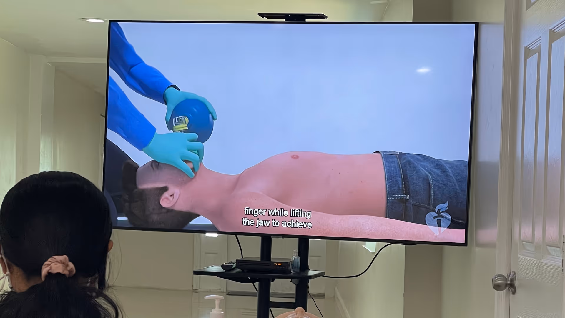 A shirtless man lies prone as gloved hands demonstrate airway technique on a TV screen.