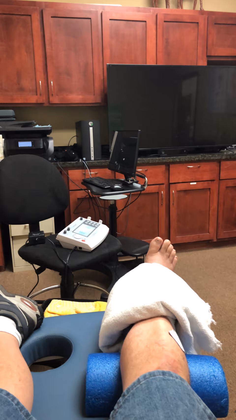 Resident's leg elevated on foam roller during therapy in a cabinet-filled room.