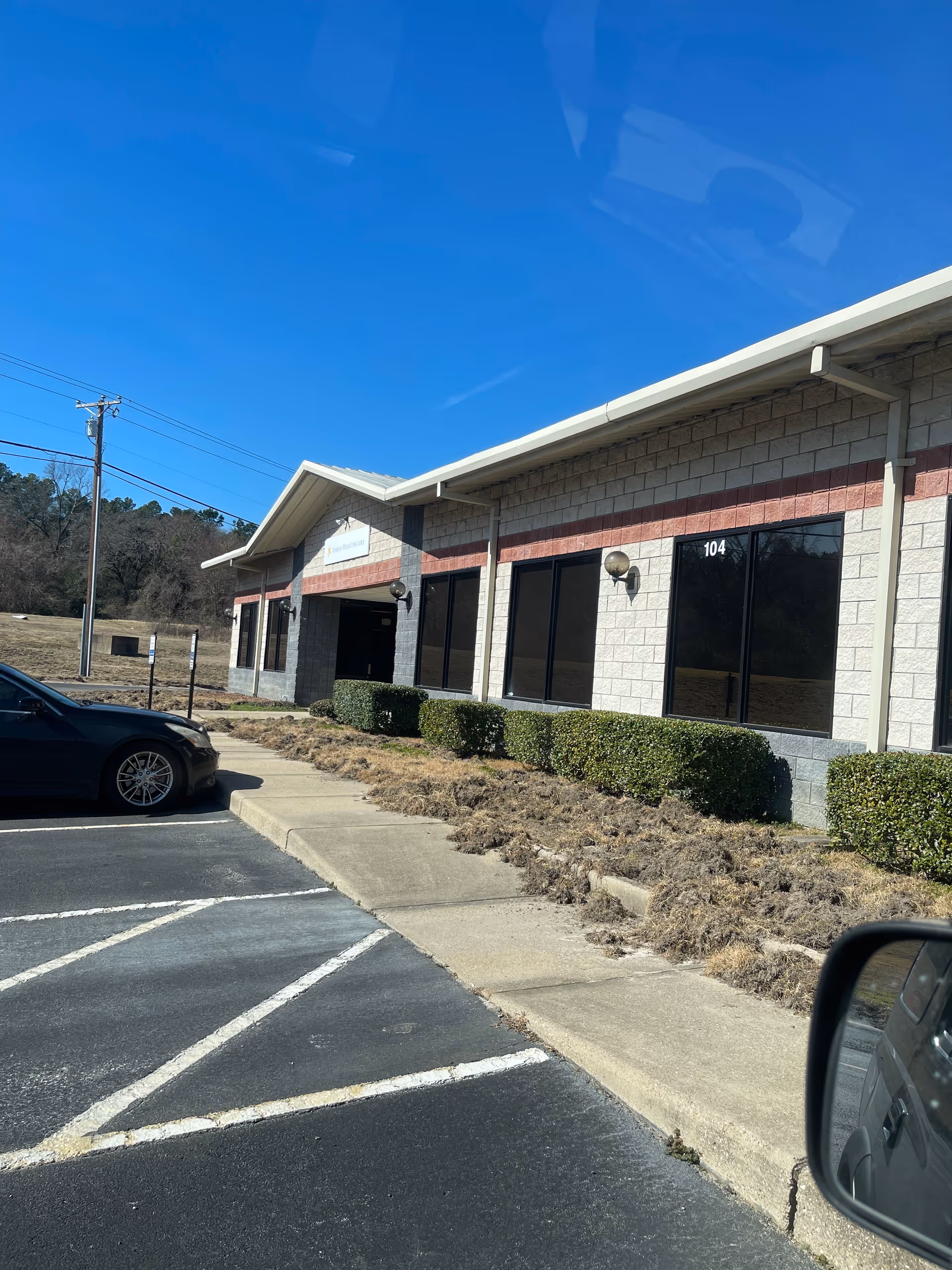 Single-story commercial building with large windows, shrubs, and a parking lot in front.