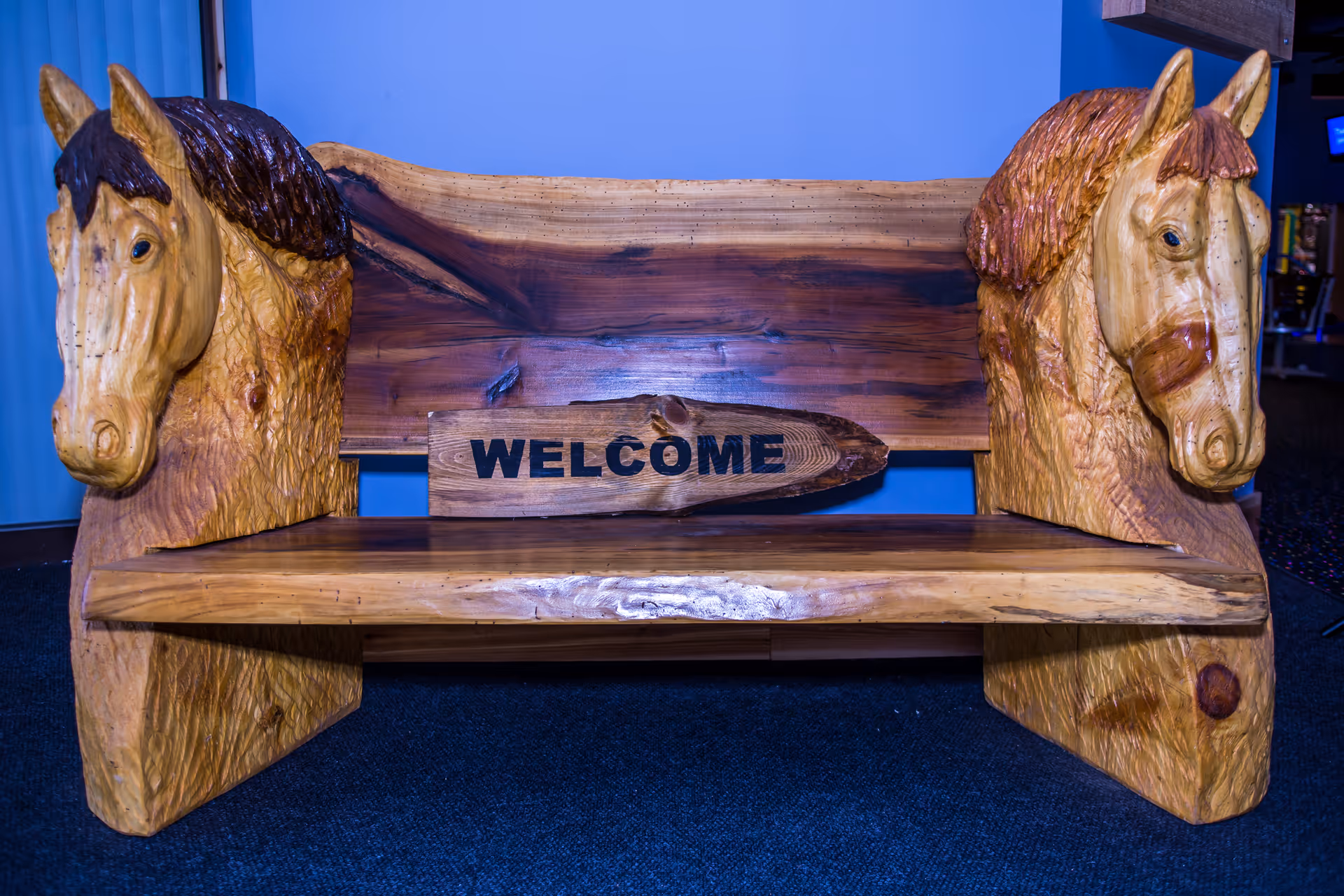 Wooden bench with carved horse heads and a welcome sign inside against a blue wall.