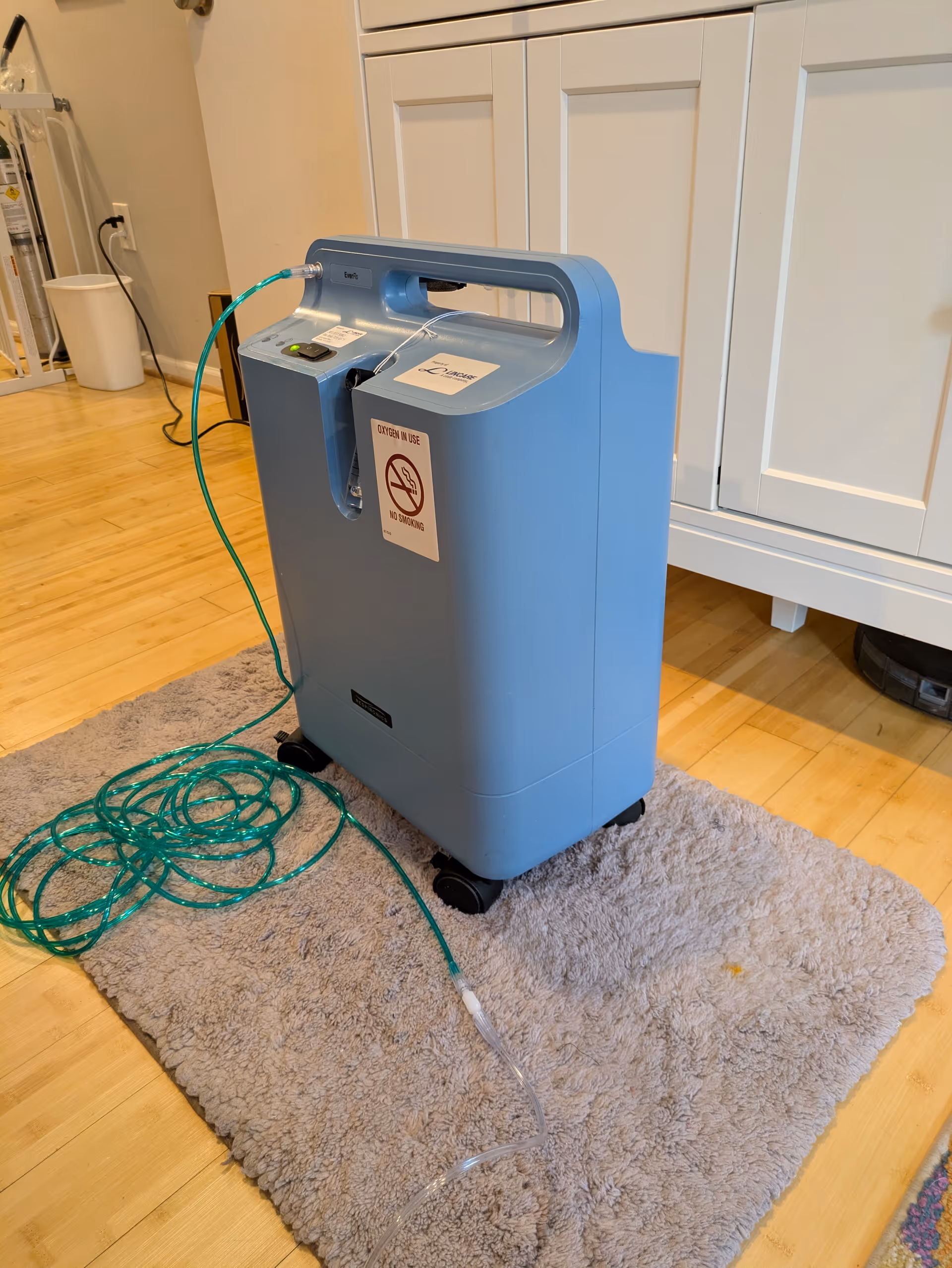 Blue LINCARE oxygen concentrator on wheels with tubing beside white cabinets indoors.