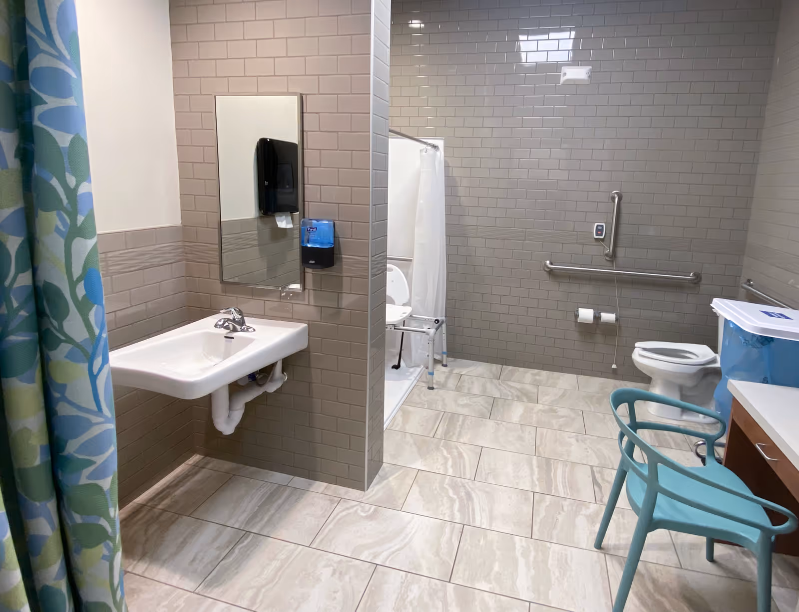 Accessible bathroom with sink, mirror, shower stall, toilet, grab bars, and aqua chair.