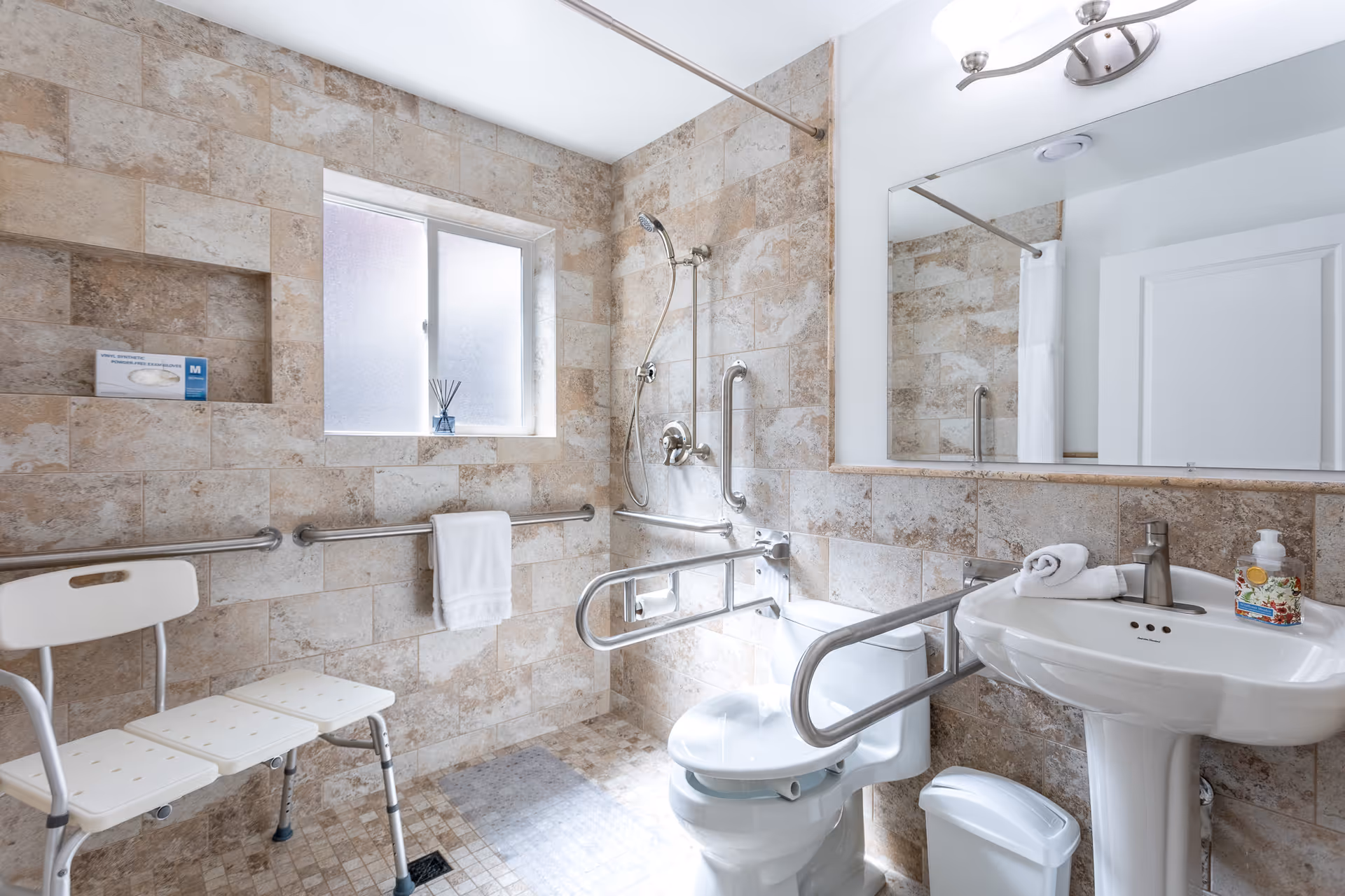 Accessible bathroom with tiled walls, grab bars, a shower, toilet, sink, and a chair.