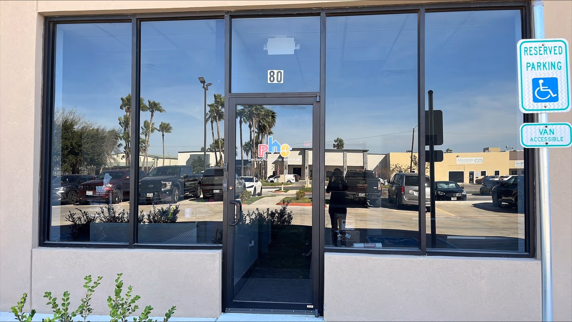 Front entrance of a building with large glass doors, parking lot, and palm trees.