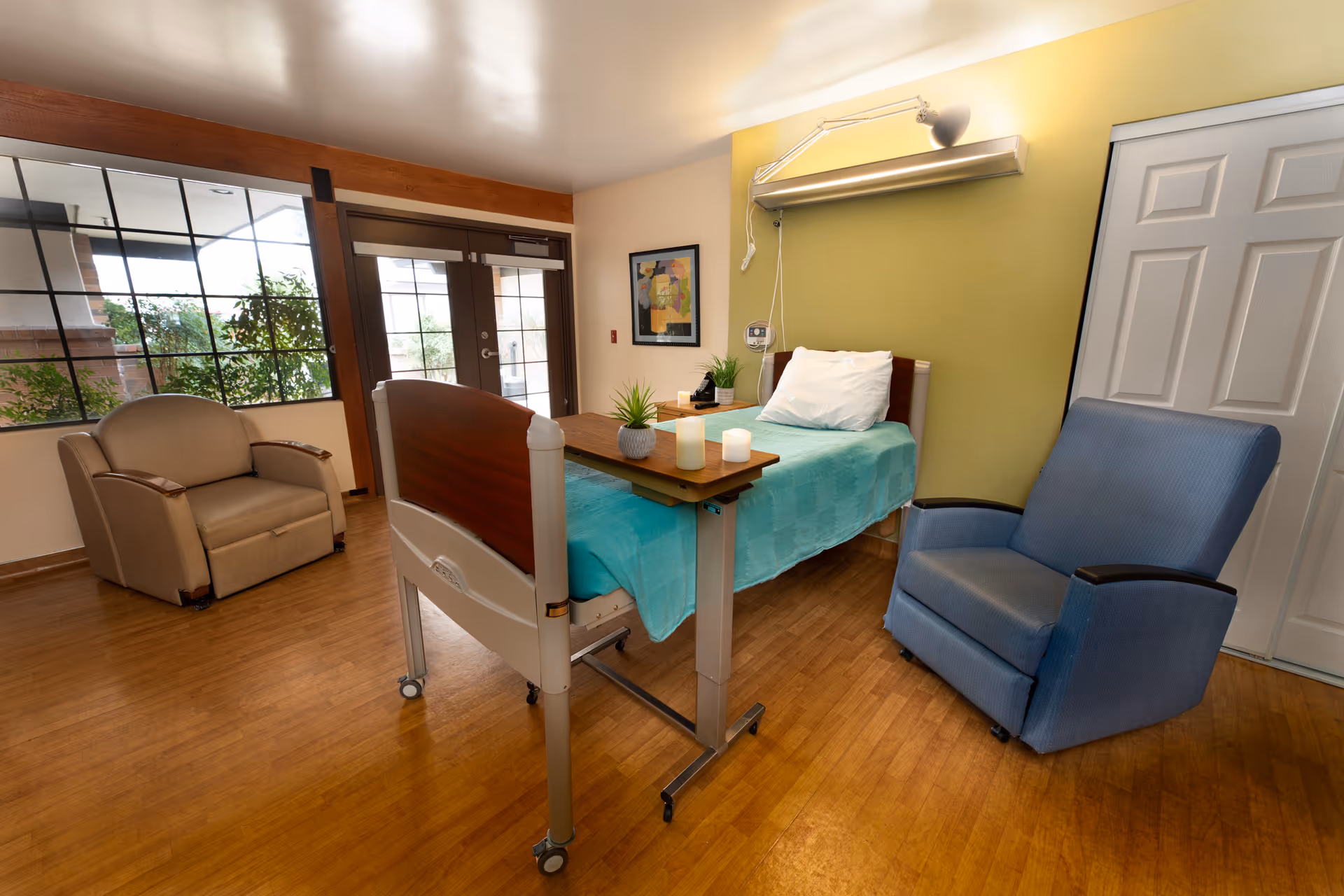 Hospice patient room with a hospital bed, teal bedspread, recliner, and glass doors.