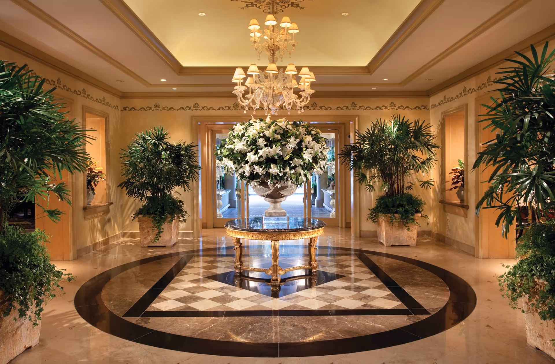 Luxurious hotel-like lobby with chandelier, large floral centerpiece, marble floor, and potted plants.