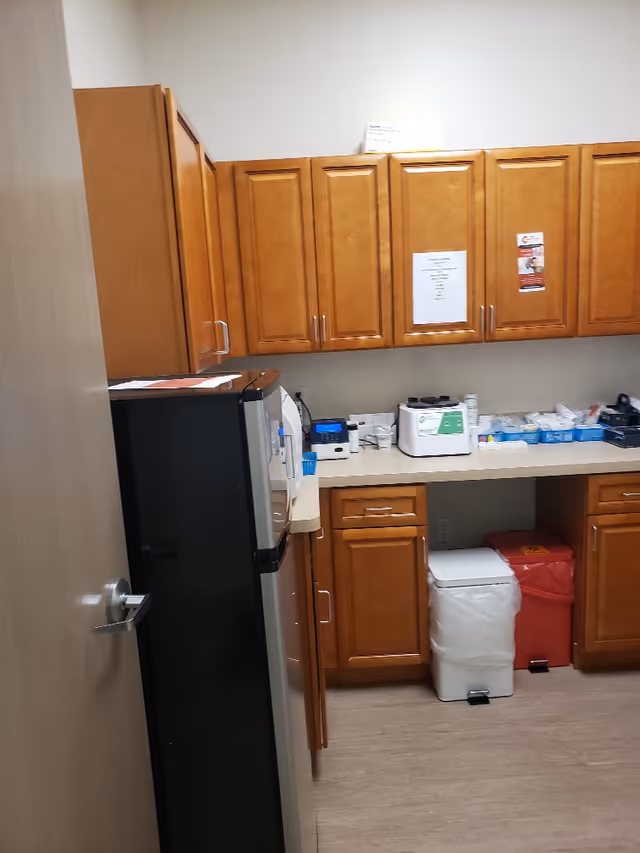 Wooden cabinets line a kitchenette with a fridge, countertop, and medical supplies in a care facility.