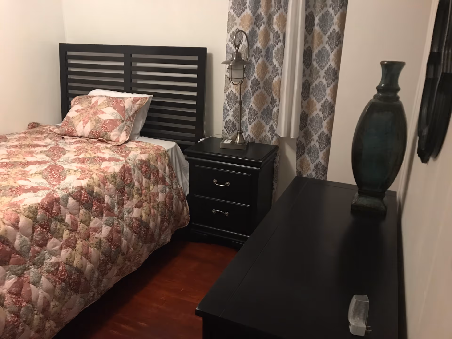 Bedroom with dark wood furniture, quilted bed, nightstand, lamp, and decorative vase.