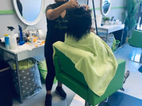 A stylist trims a client's hair in a green chair inside a salon.