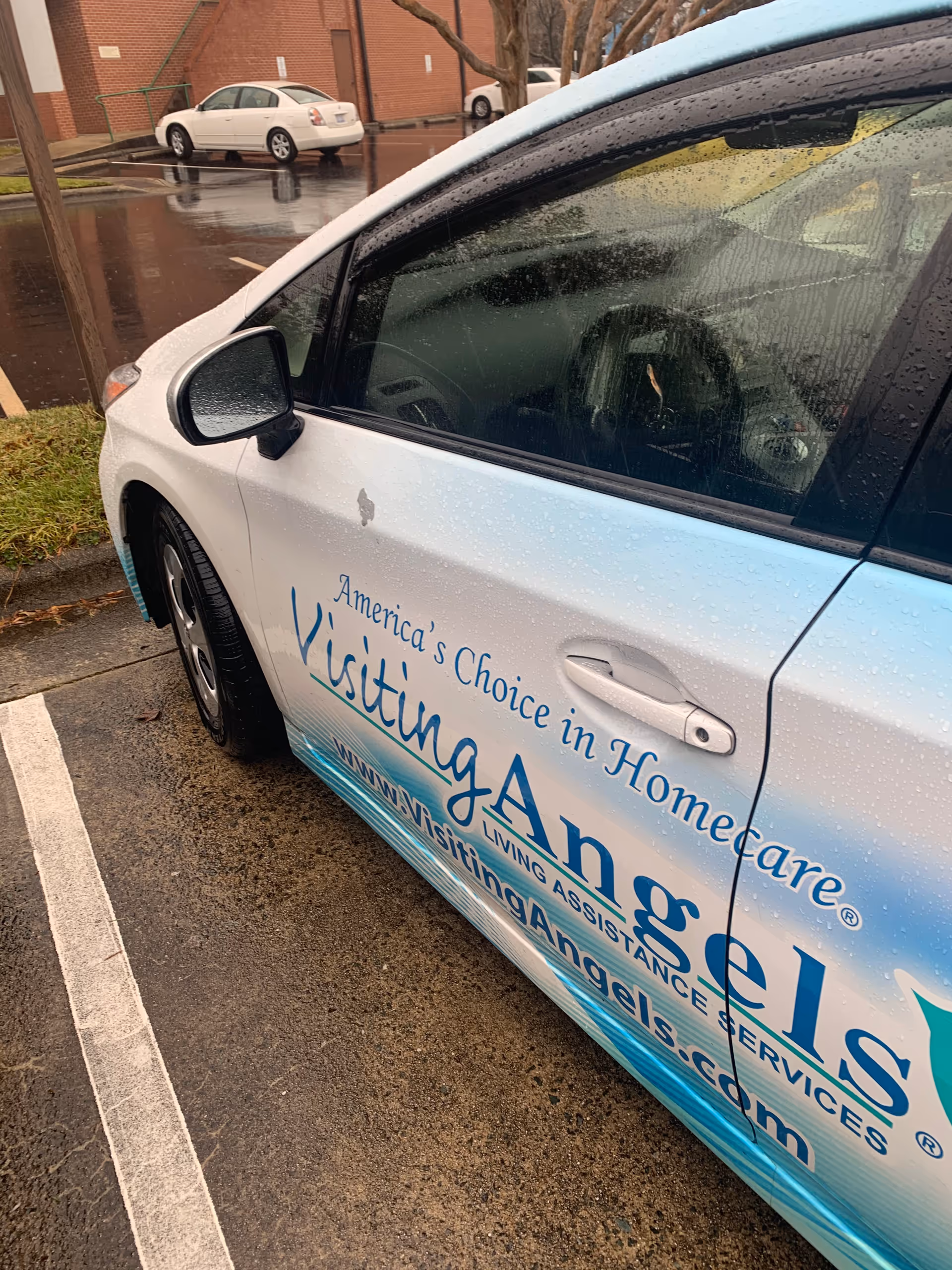 White Visiting Angels van parked in a rainy lot beside a brick building.