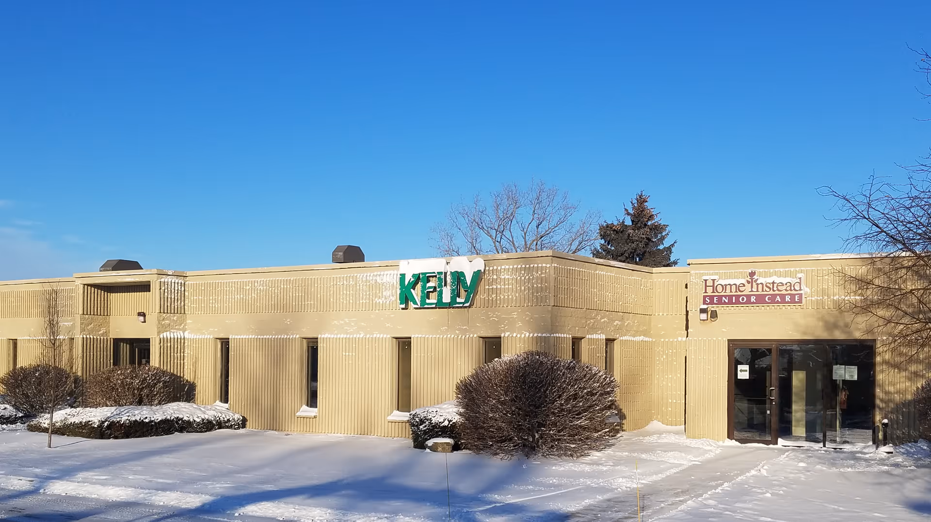Exterior view of a beige senior care building with Home Instead sign and snow on ground.