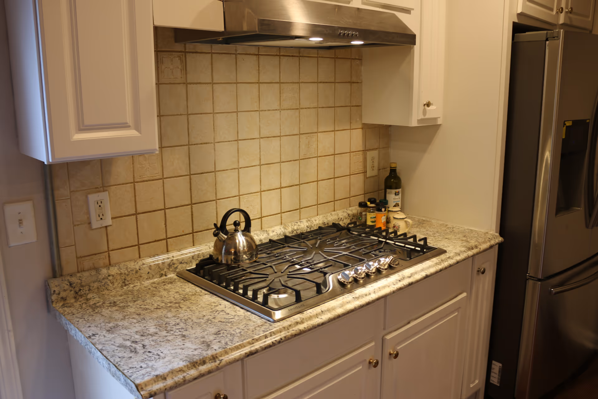 Stainless steel gas cooktop with kettle, granite counter, and refrigerator.