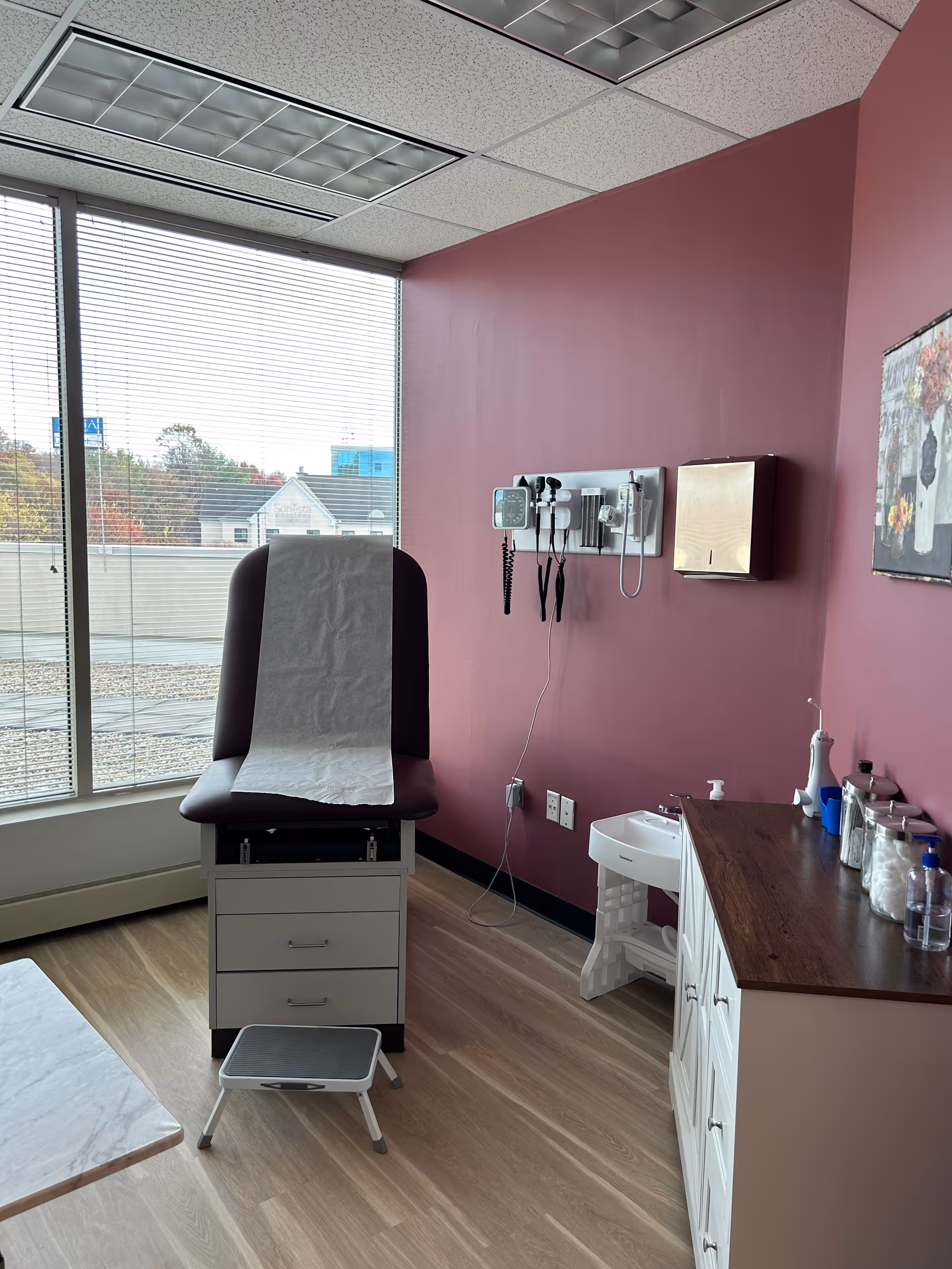 Medical examination chair and supplies in a clinic room with burgundy walls and a large window.