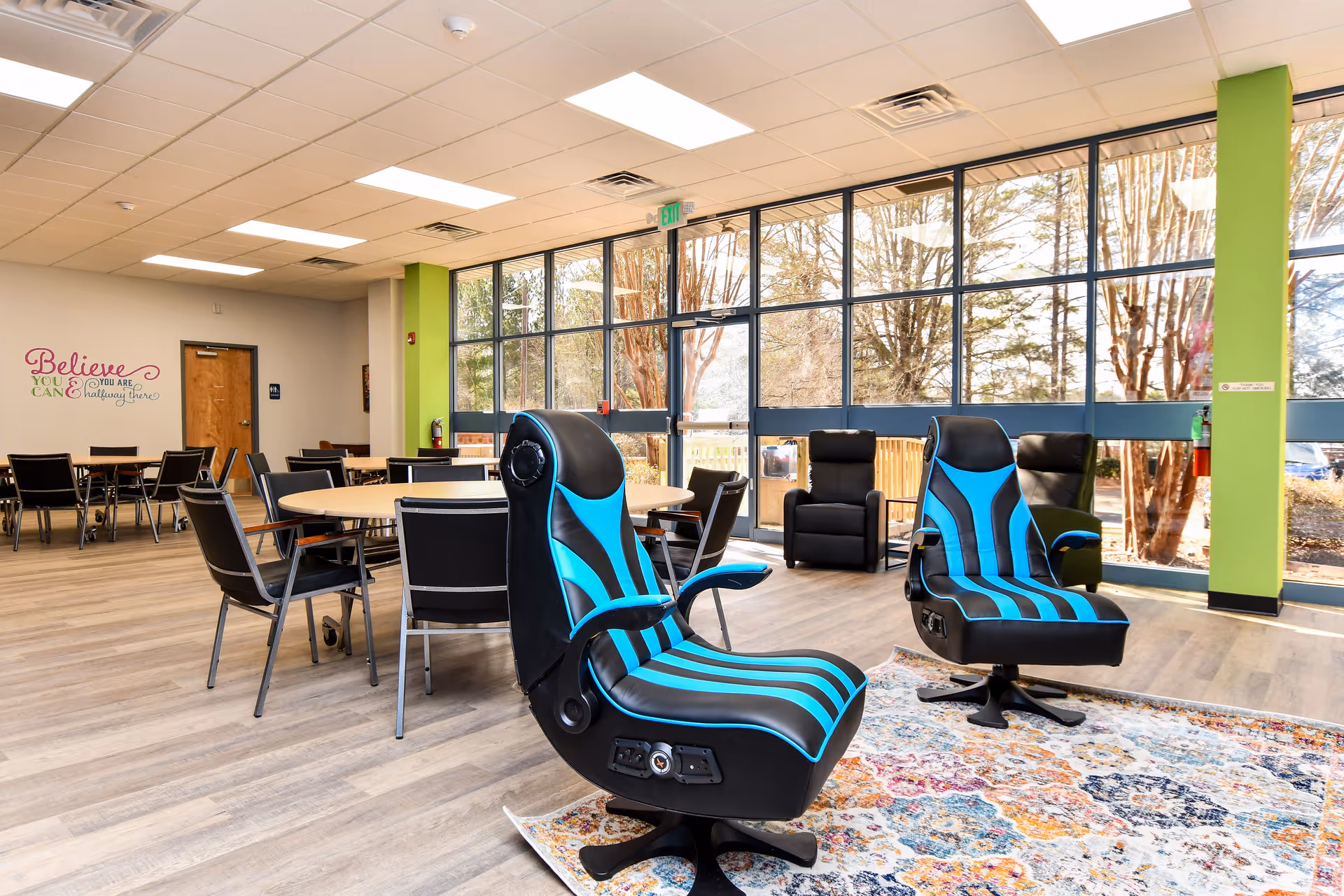 Bright senior living lounge with large windows, round tables, and blue-accent recliners.