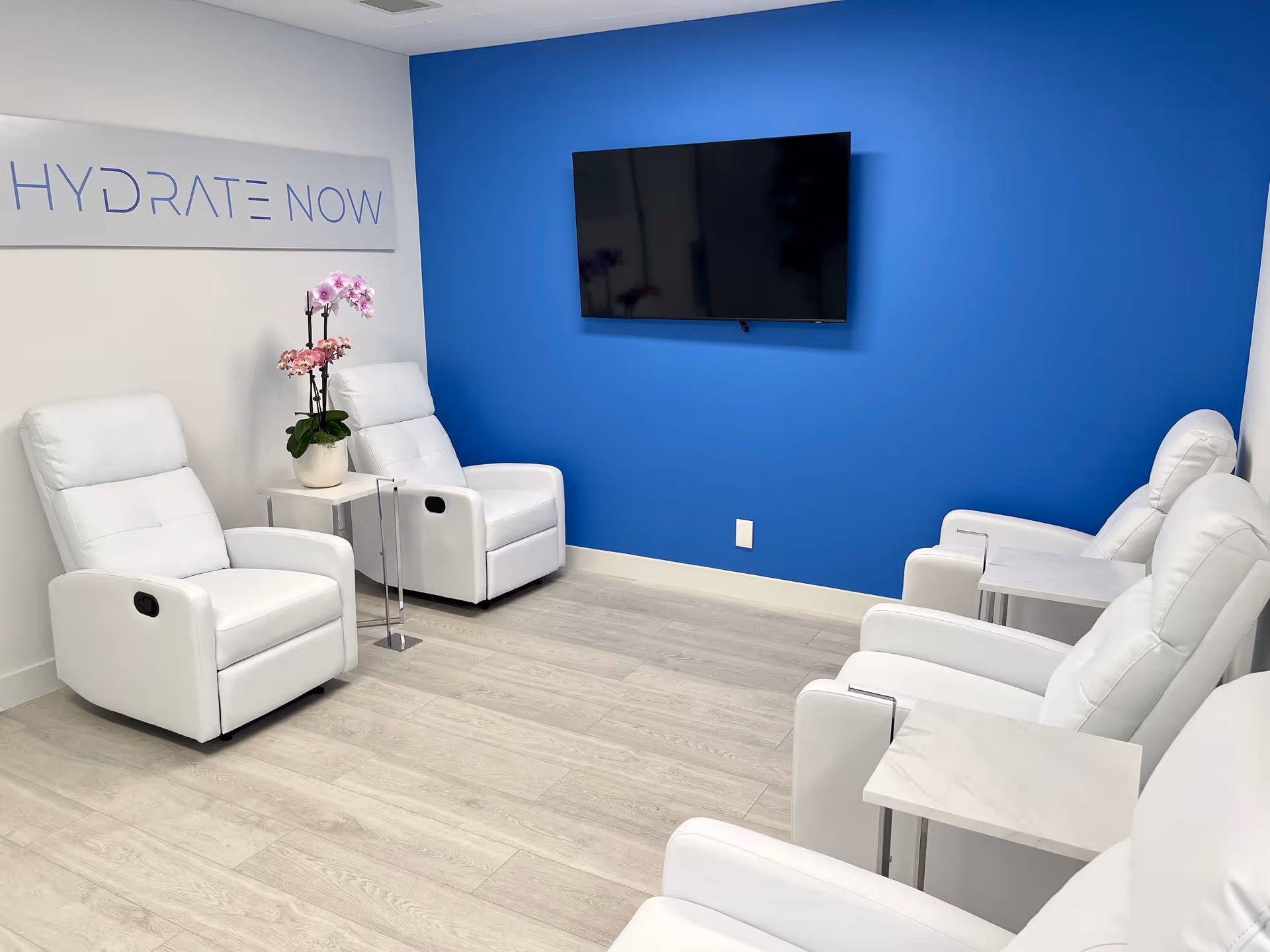 A bright white lounge area with white chairs, Hydrate Now sign, blue wall, mounted TV.
